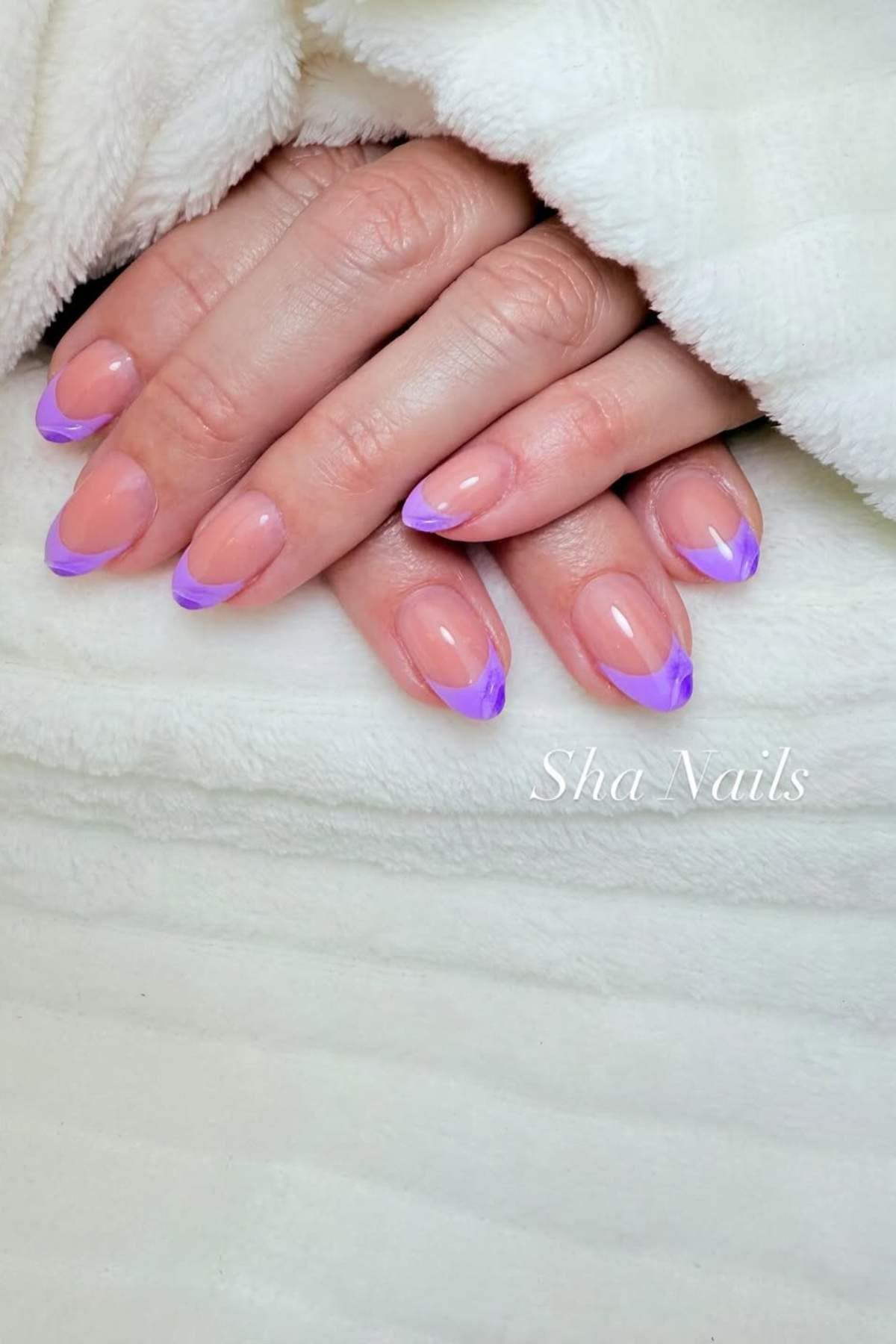 A hand with almond-shaped nails featuring a nude base and pastel purple French tip design rests on a white textured blanket.