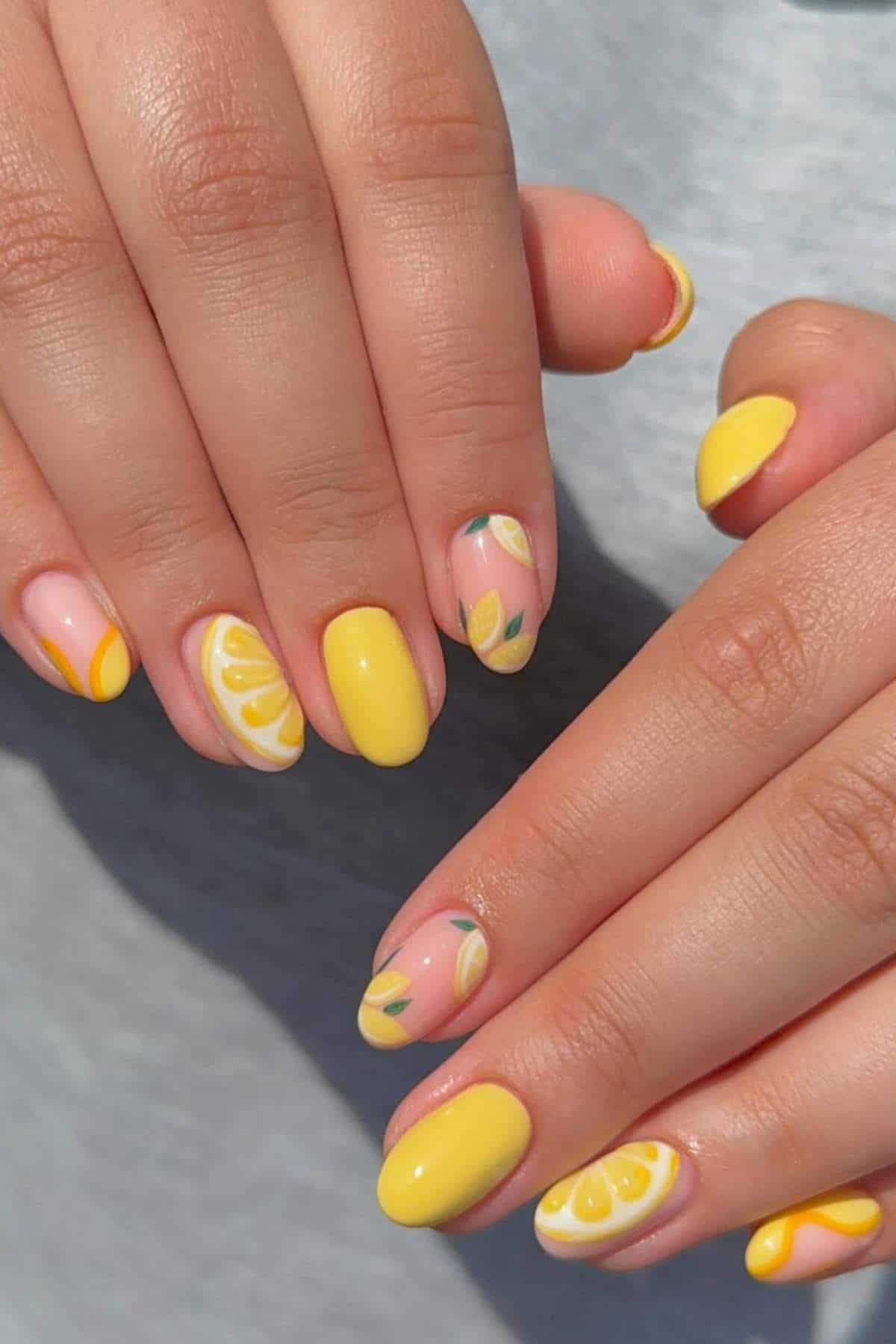 Hands with manicured nails featuring bright yellow polish and lemon slice designs on a light gray background.