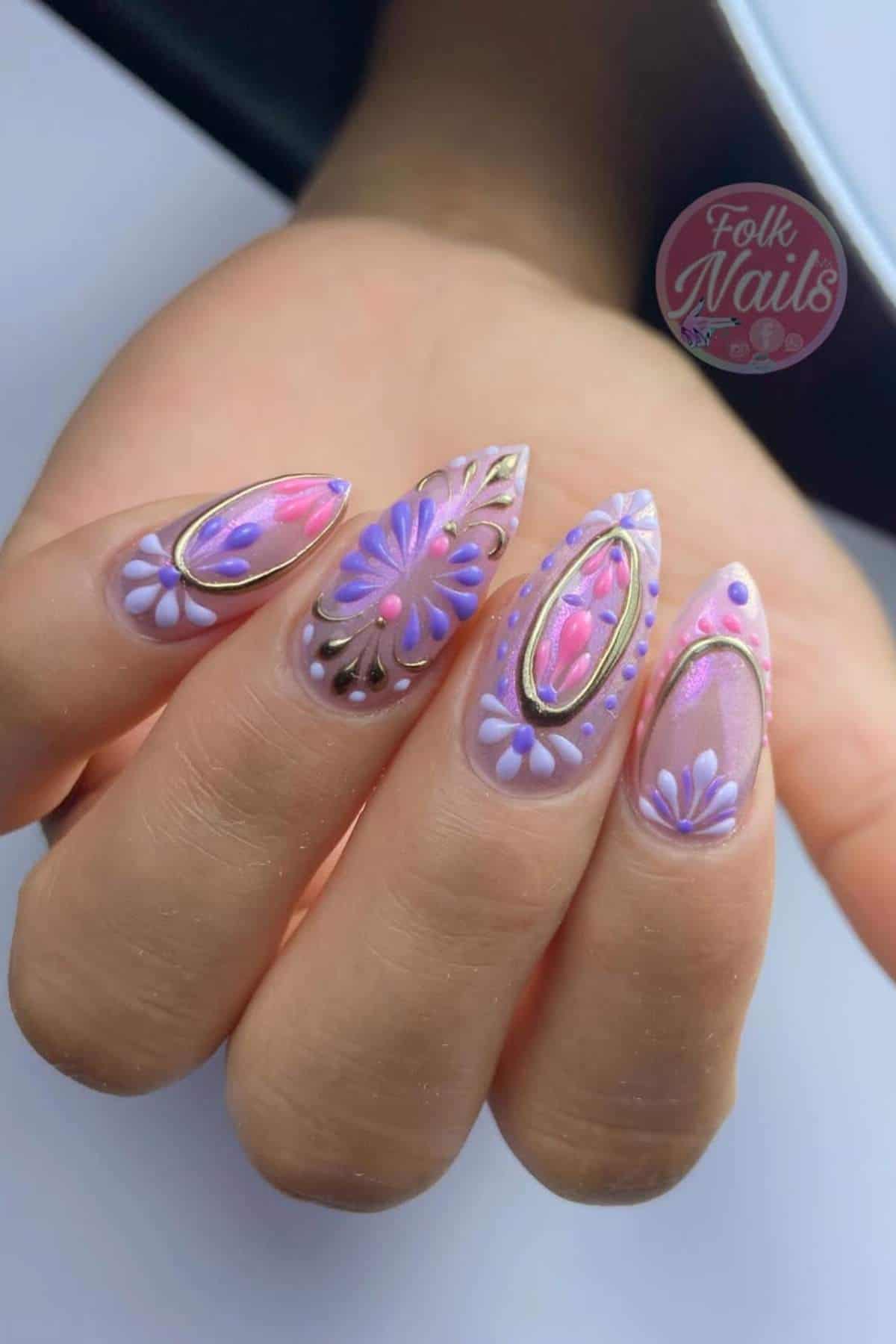 A hand with long, almond-shaped nails featuring 3D purple and pink floral designs, gold accents, and intricate details on a light base.