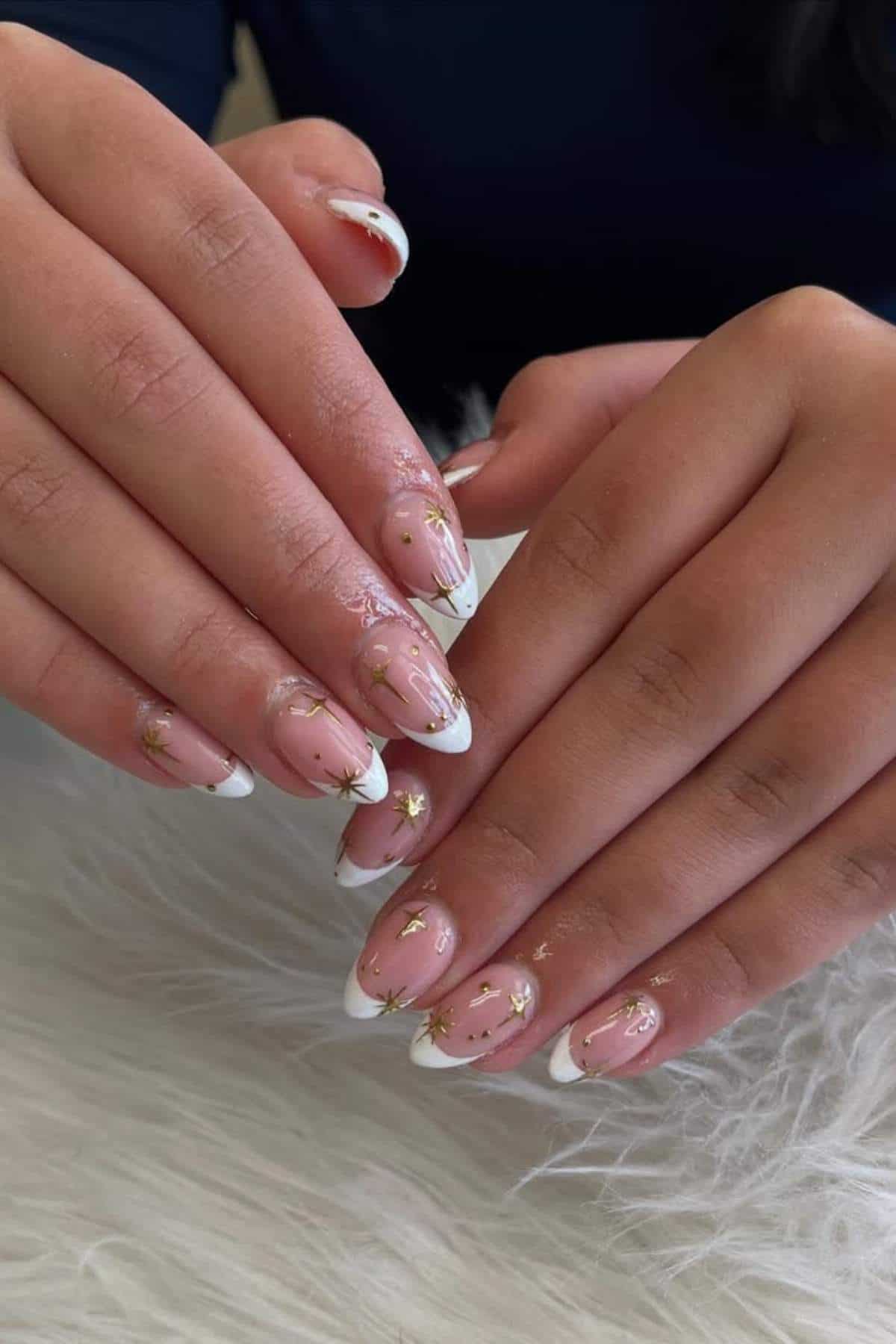 Hands with almond-shaped nails featuring a white French tip design, gold foil accents, and subtle nail art, resting on a white textured surface.