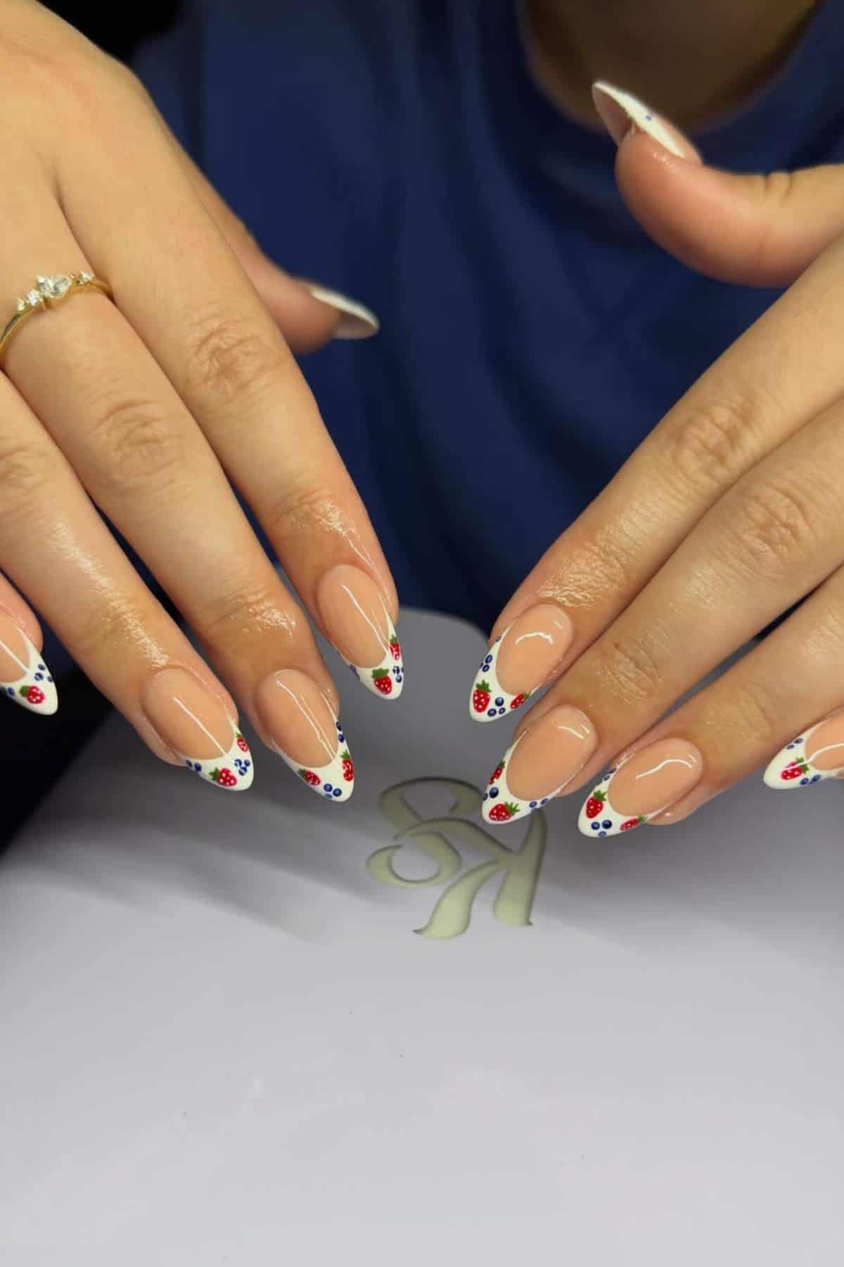 Hands with almond-shaped nails featuring a nude base and white tips decorated with small red, green, and black floral designs. A gold ring with pearls is worn on one finger.