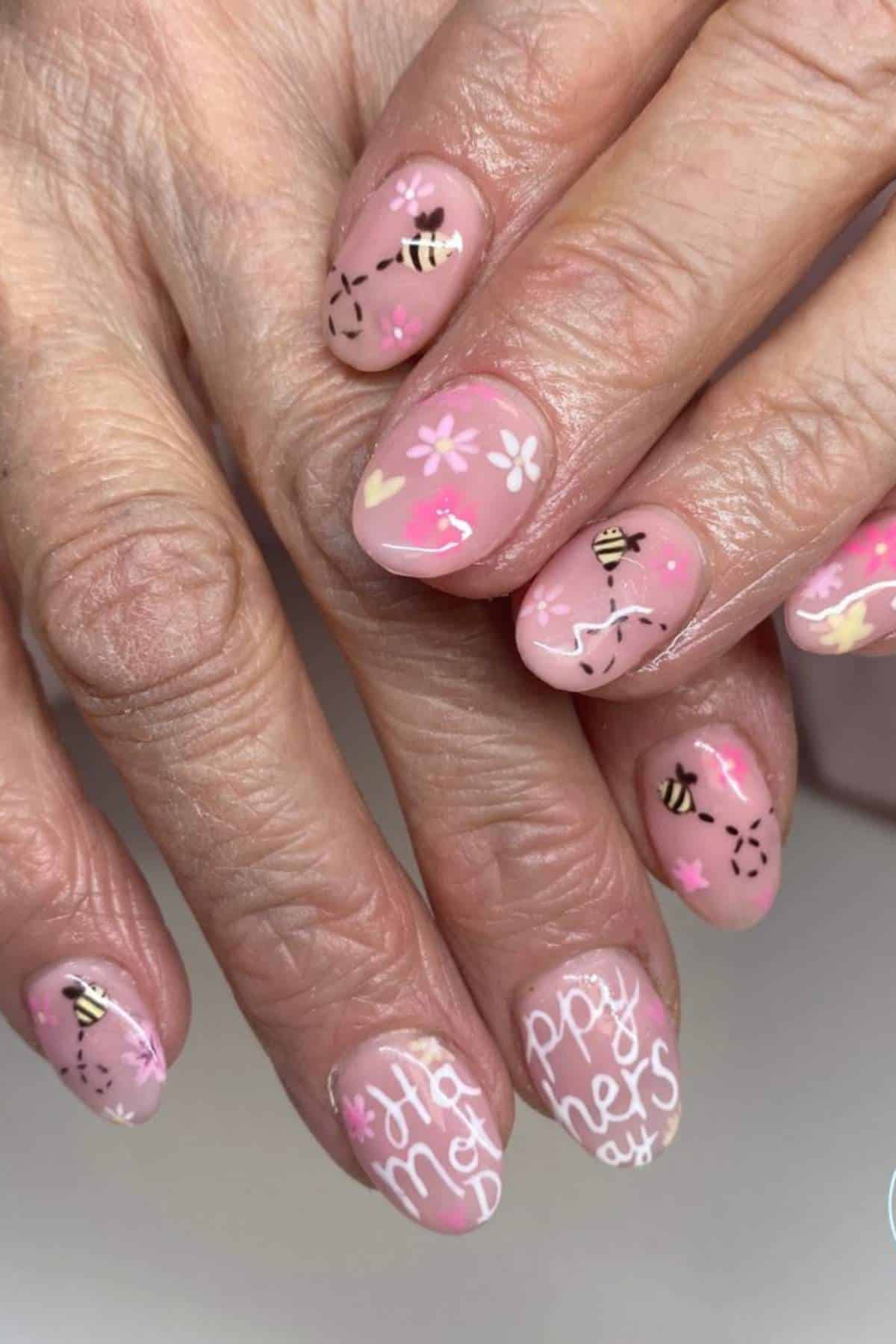 A hand with light pink manicured nails featuring bee, flower, and heart designs, and “Happy Mother’s Day” written on the ring finger.