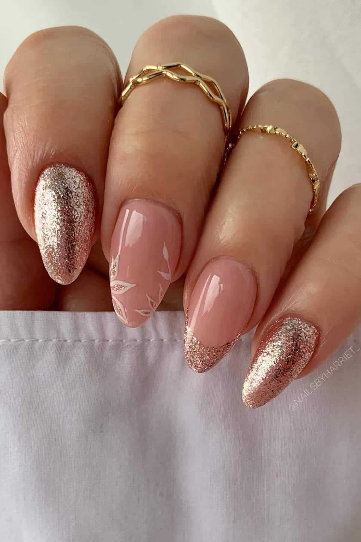 A hand with almond-shaped nails featuring pink polish, glittery rose gold tips, and a white leaf design on one nail; two gold rings are worn on the fingers.