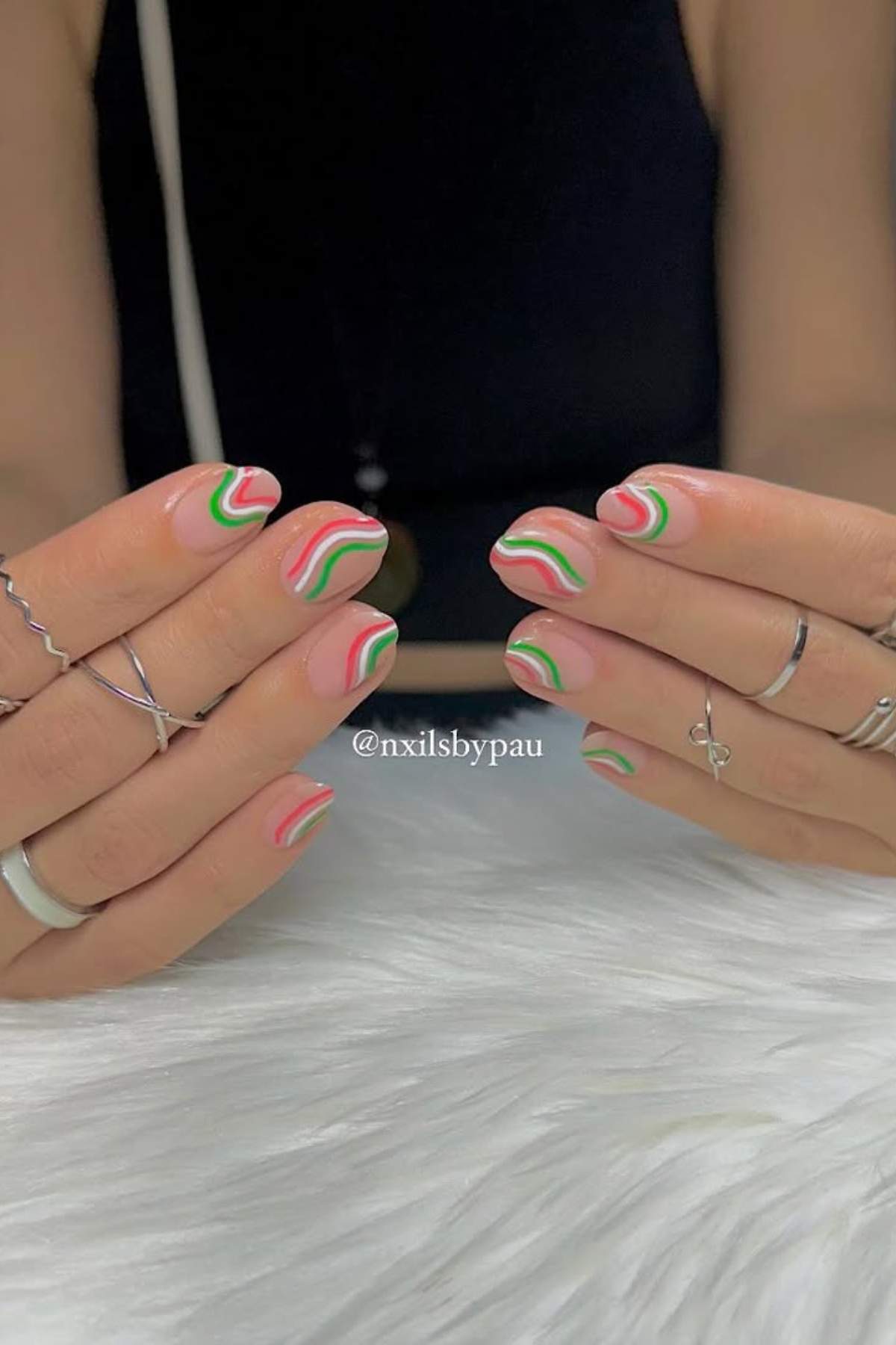 Hands with short nails featuring wavy pink, red, and green designs on a nude base. The person is wearing multiple silver rings and has their hands positioned over a white surface.