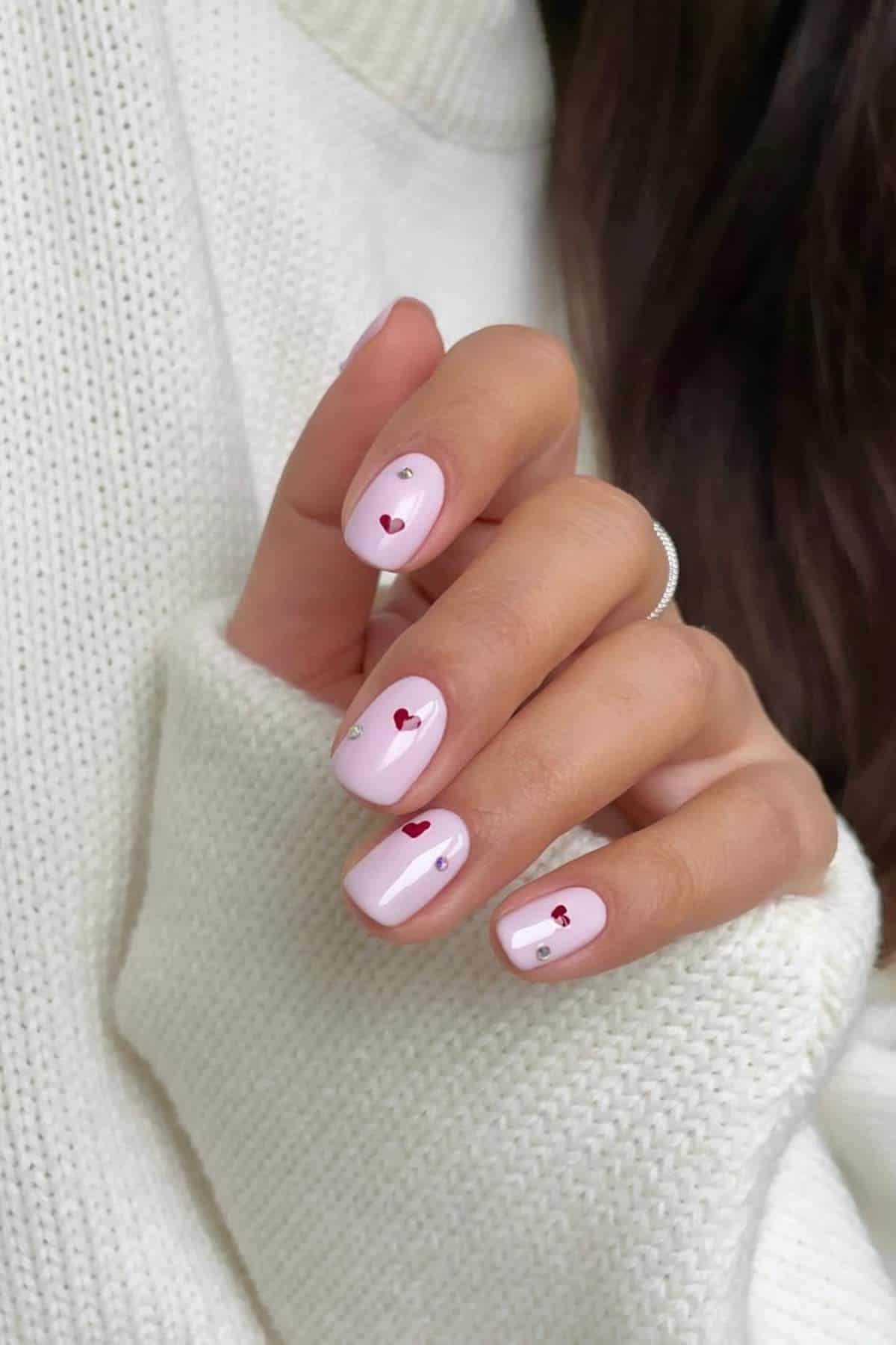 A hand with light pink nail polish, each nail decorated with a small red heart and a tiny silver gem, against a white sweater background.