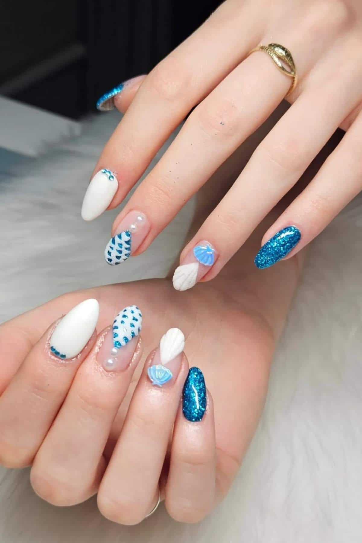 Hands with almond-shaped nails featuring blue and white designs, including glitter, patterns, pearls, and 3D embellishments, displayed on a white textured surface.