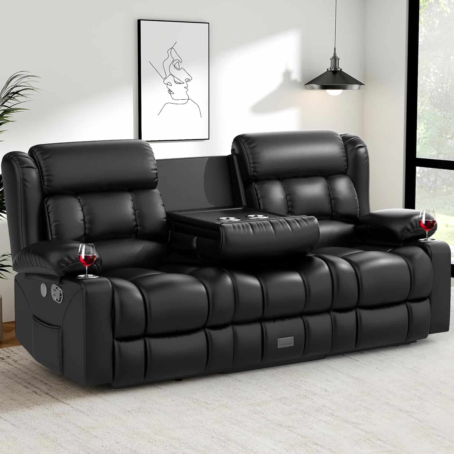 A black leather reclining sofa with a fold-down center console holding two wine glasses, placed in a modern living room with wall art and a floor-to-ceiling window.