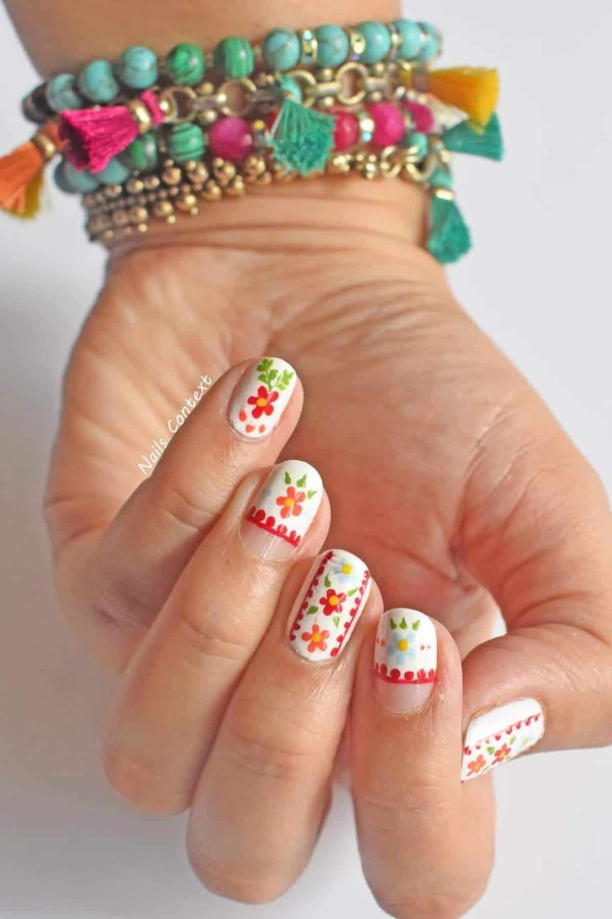 A hand with white nail polish featuring colorful floral and dotted designs, wearing beaded bracelets with tassels in various colors.