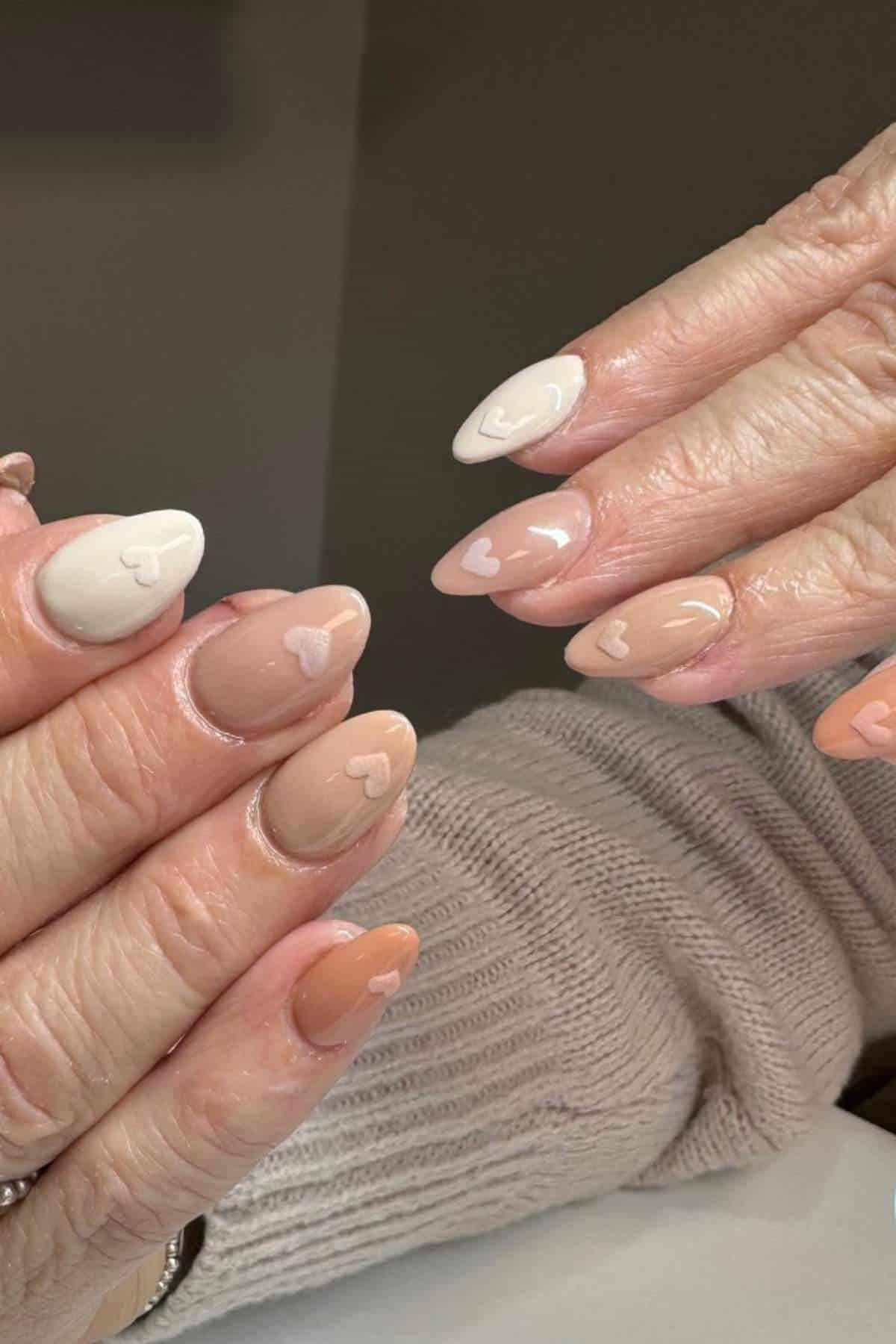 Hands with almond-shaped nails painted in neutral and peach tones, each nail featuring a small, simple heart design near the tip.
