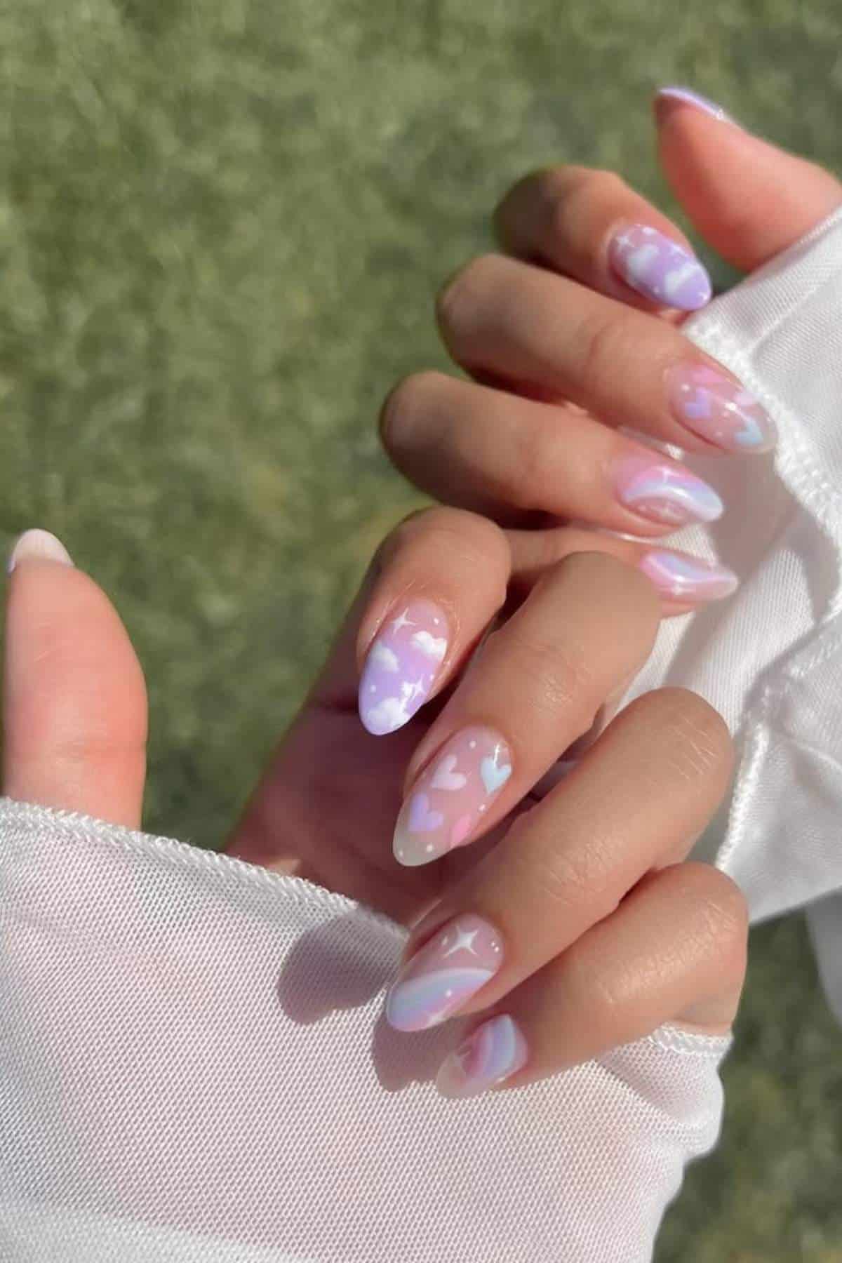 A hand with almond-shaped nails features pastel pink and purple nail art with heart and swirl designs, shown against a blurred green background.