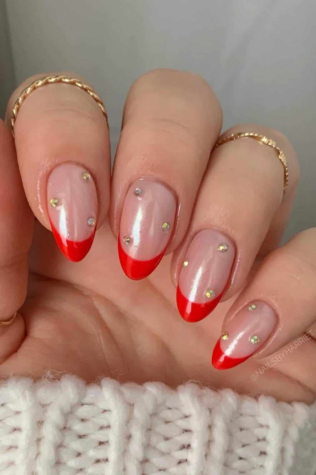 A hand with almond-shaped nails featuring a nude base, red tips, and small rhinestone accents, wearing gold rings and a white knit sleeve.