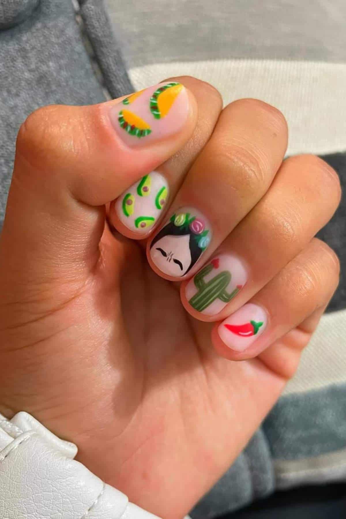 A hand with colorful nail art, including designs of avocados, a woman's face, a cactus, a chili pepper, and a lemon slice.