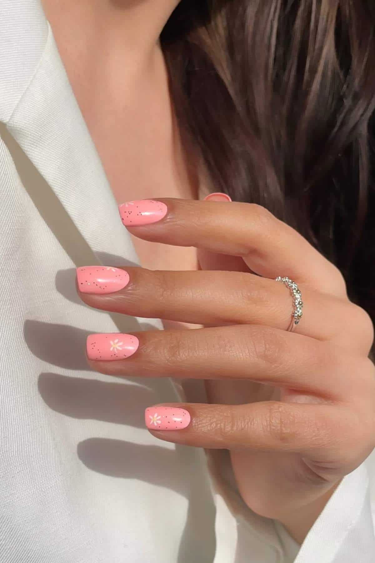 A hand with pink, glossy, square-shaped nails featuring small white flower designs and glitter, wearing a silver ring, against a white fabric background.