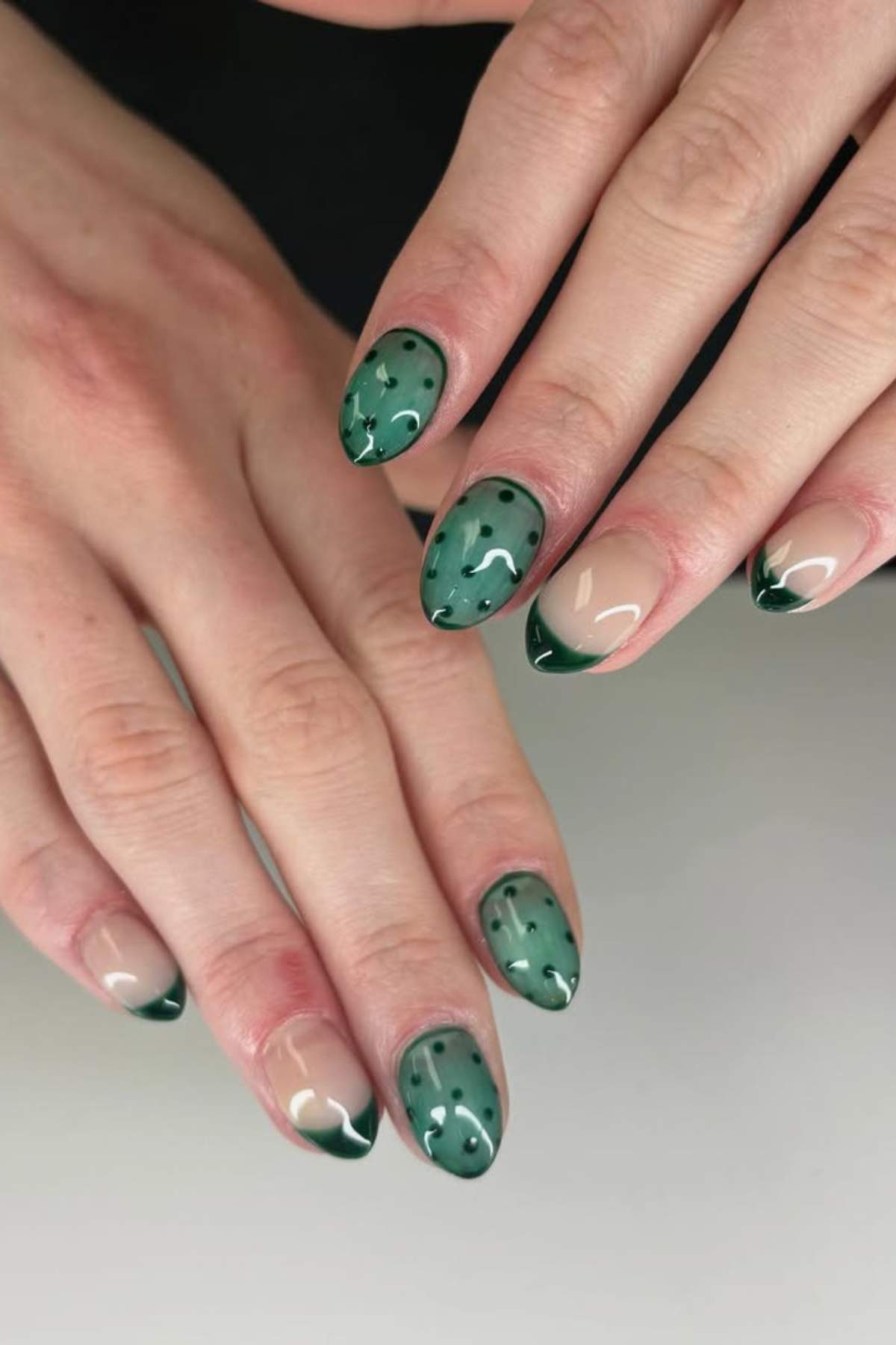A close-up of hands with almond-shaped nails featuring green and nude polish; some nails have a cactus design, others have a green French tip.