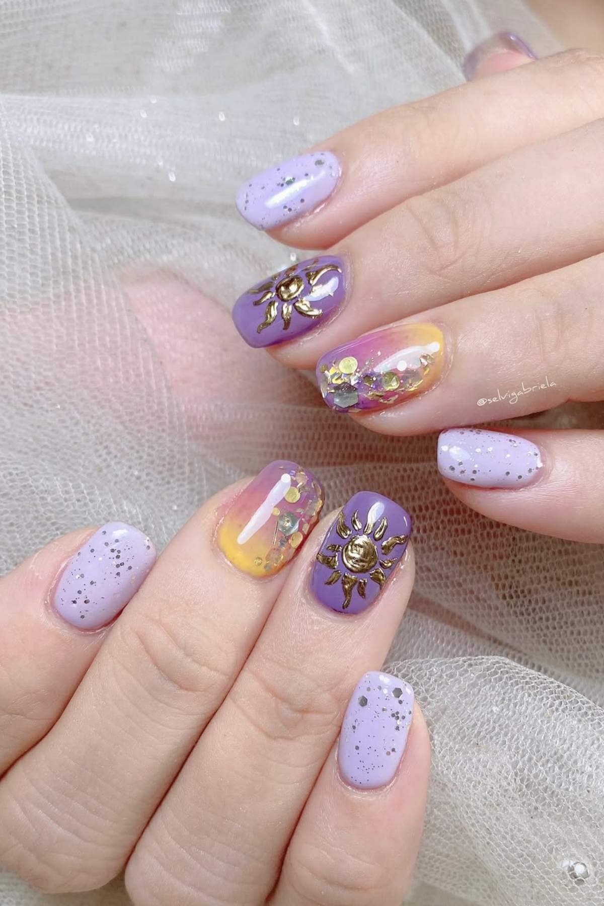 Hands with lavender and yellow gradient nails, sparkly accents, sun-shaped gold embellishments, and glitter details on a soft fabric background.