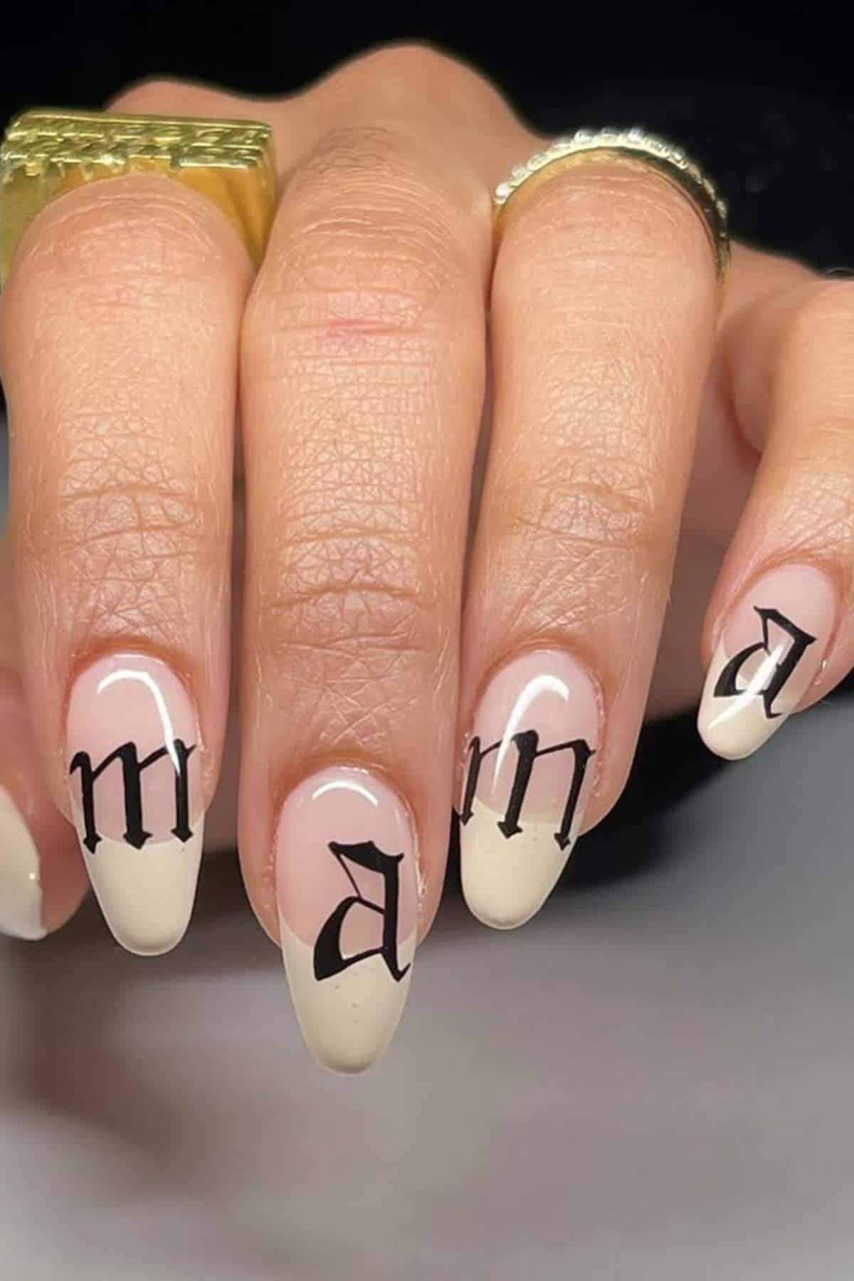 A hand with almond-shaped nails painted in a nude and white design, each nail featuring a single black letter in Gothic script.