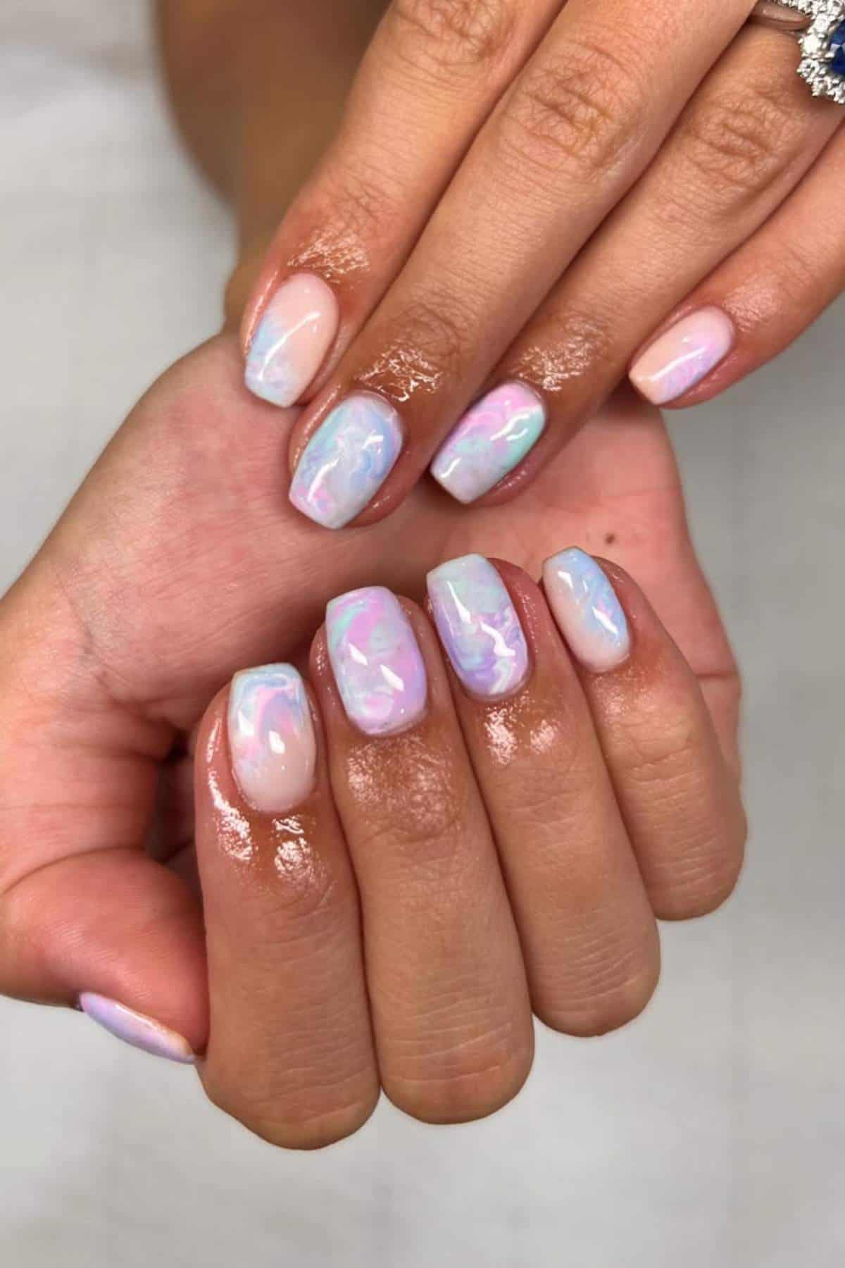 Hands with pastel, iridescent marble nail art on short, square-shaped nails. One hand has a diamond ring on the ring finger.