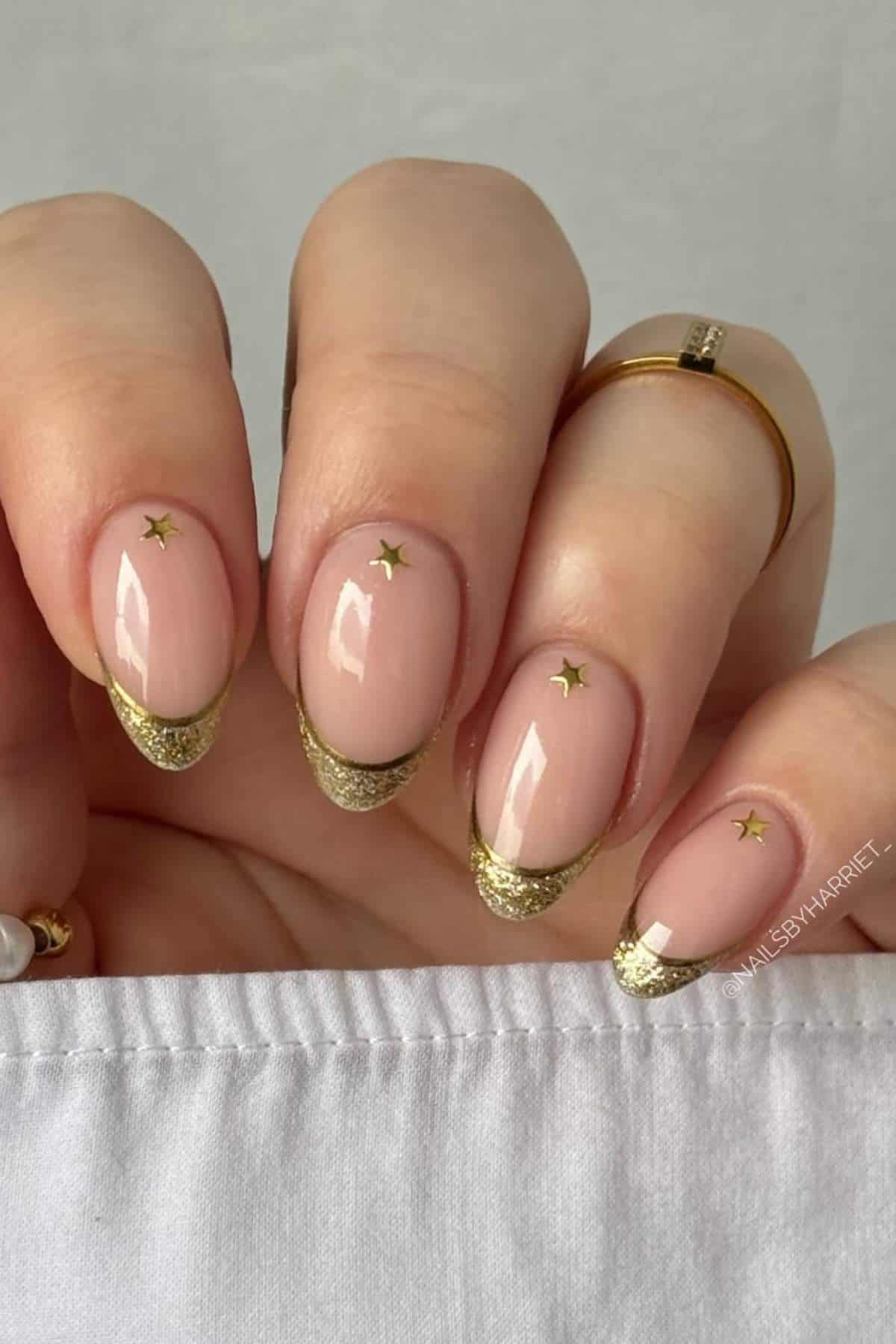 A hand with oval-shaped nails features nude polish, gold glitter French tips, and small gold star decals; the person wears simple gold rings.