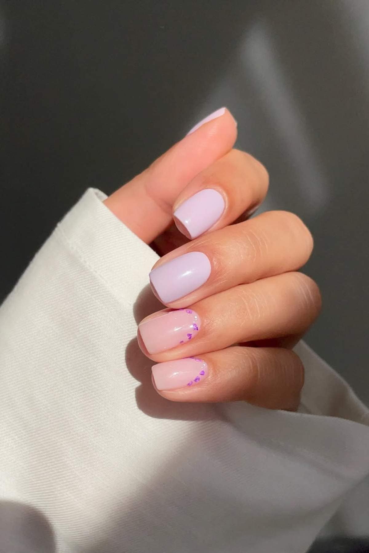 A hand with short, square-shaped nails painted light pink; two nails have small purple dot accents near the tips. The hand is partially covered by a white long sleeve.
