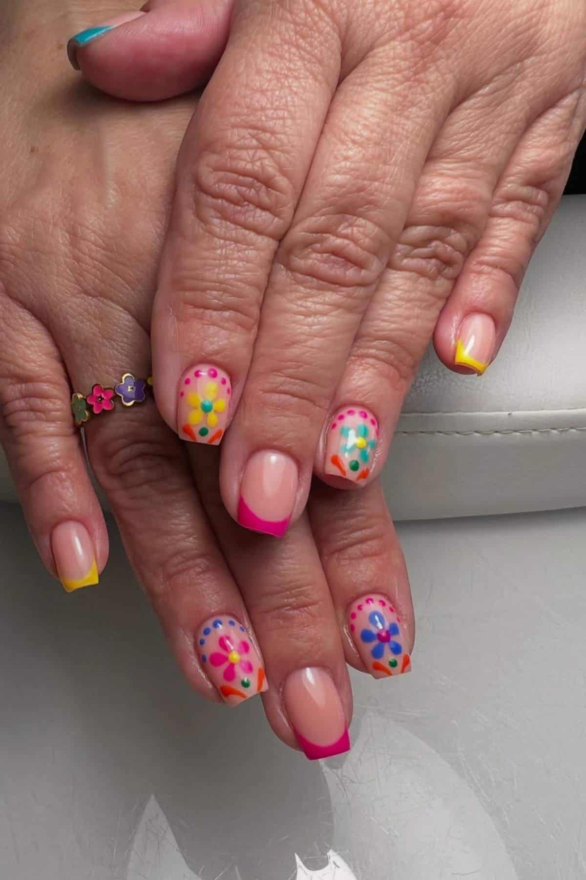 Hands with brightly colored French manicured nails, featuring floral and polka dot designs in blue, pink, orange, and yellow. A floral ring is worn on one finger.