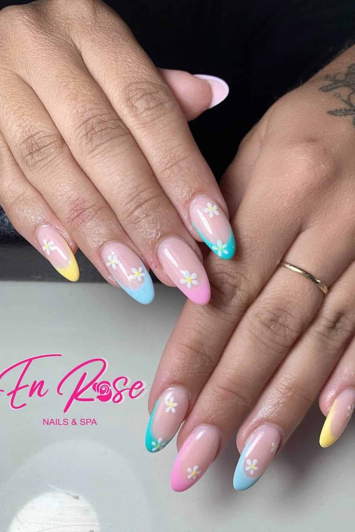 A hand with long almond-shaped nails featuring pastel yellow, pink, and blue French tips and small white daisy flower designs on each nail. The En Rose Nails & Spa logo is visible.