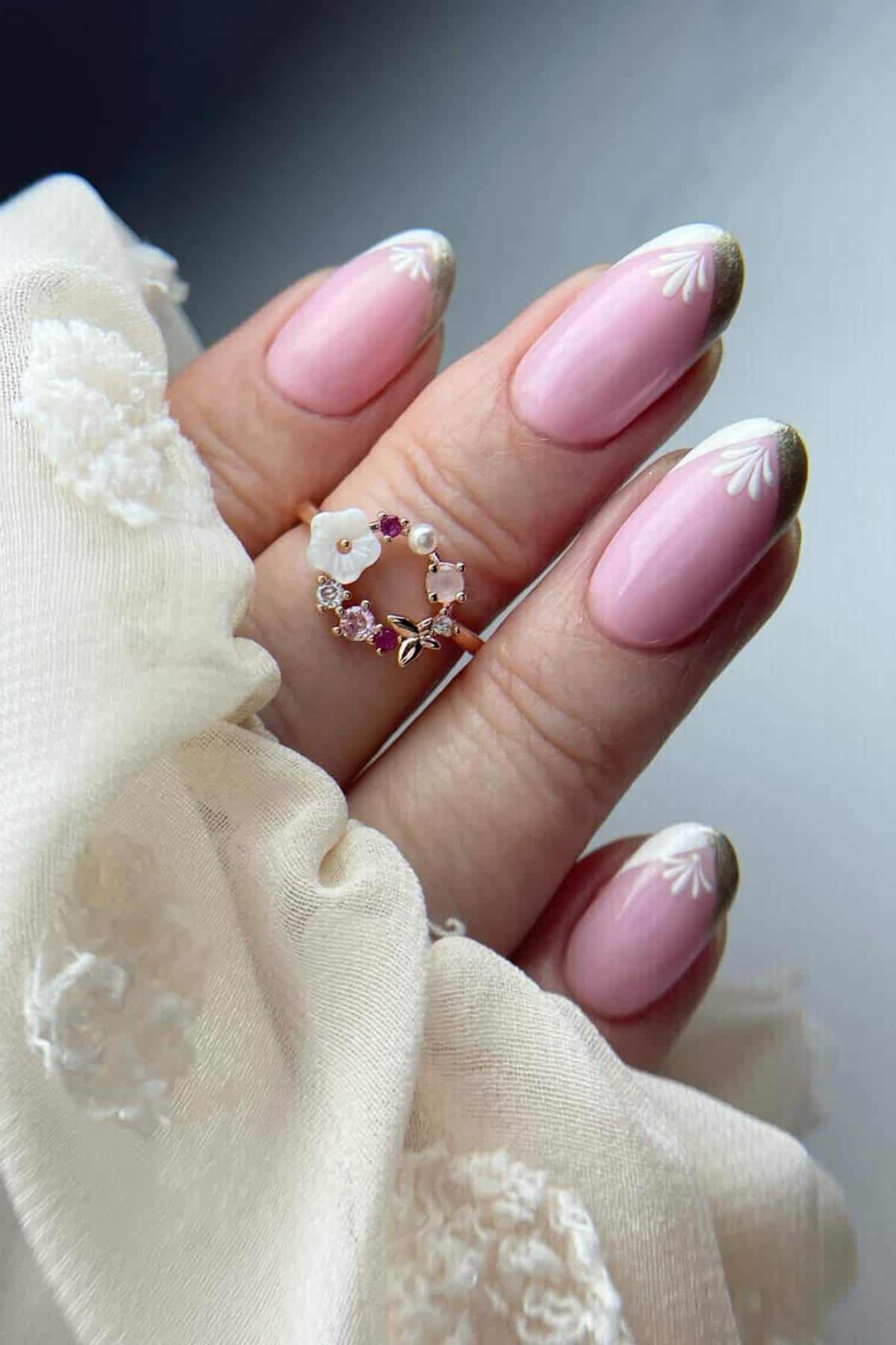 A hand with pink manicured nails featuring white and gold floral tips, wearing a gold ring with small gemstones and a flower detail, partially wrapped in a cream fabric.