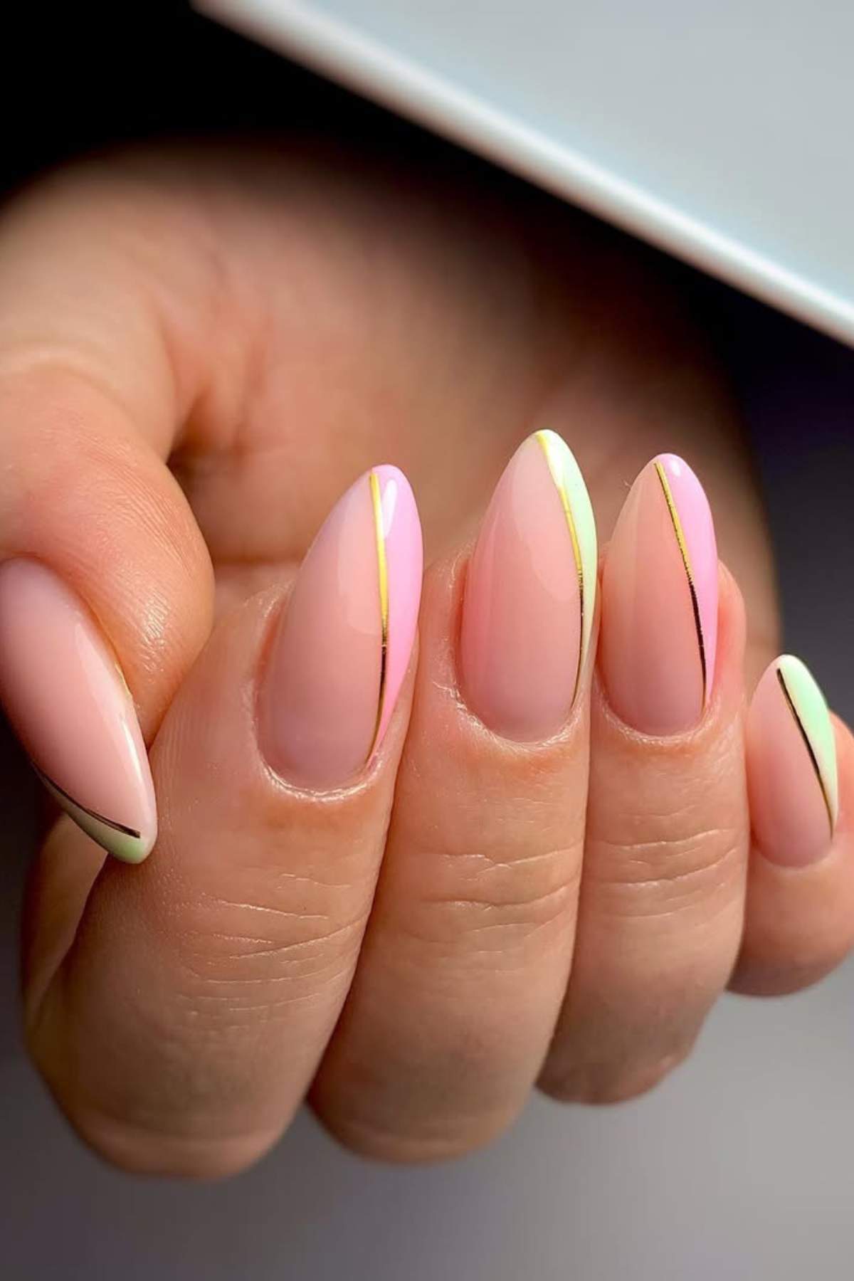 A hand with almond-shaped nails painted in a pale pink base, accented with thin gold and black diagonal lines and subtle hints of pastel colors.