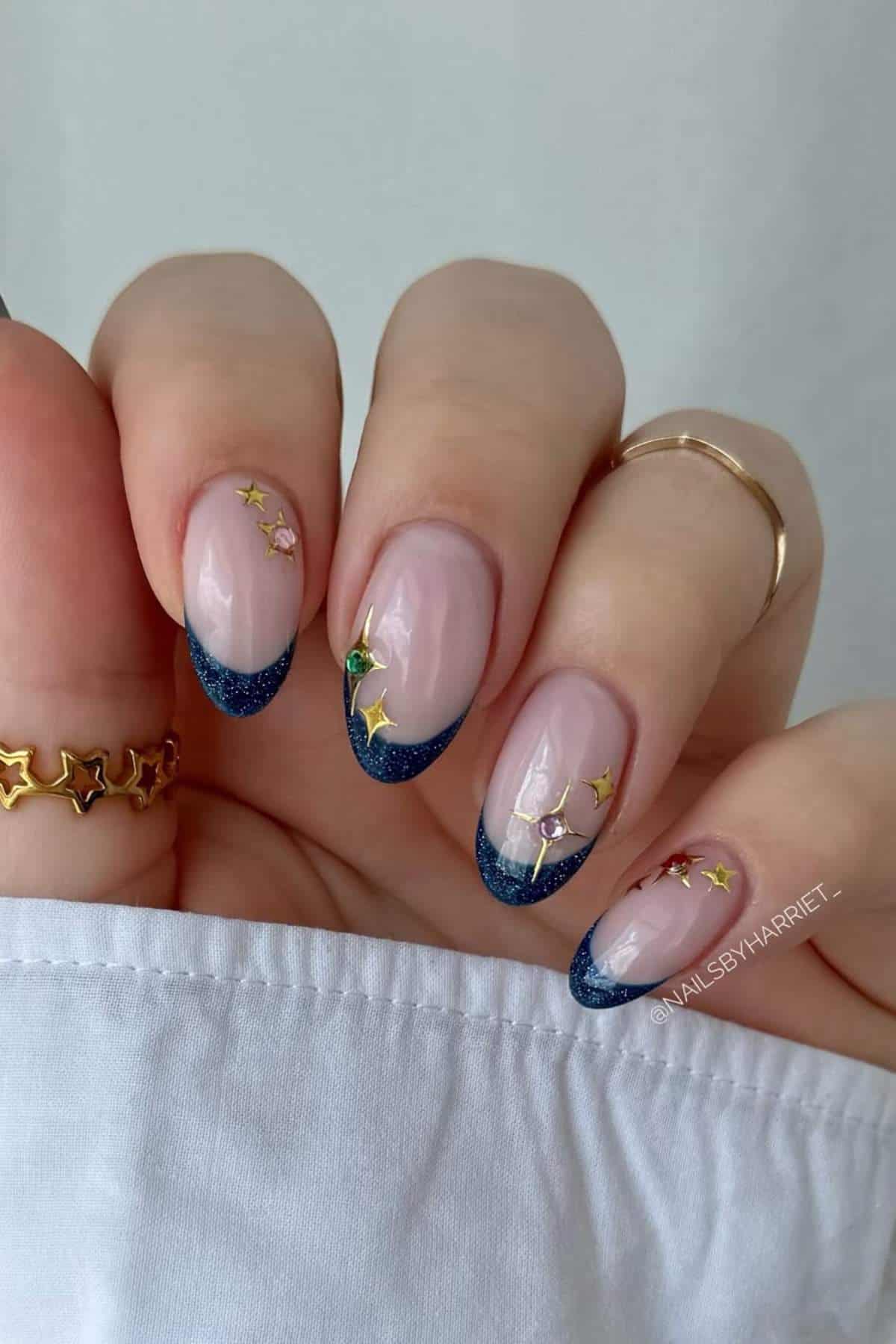 A hand with navy blue French tip nails, decorated with small gold star and butterfly decals, wearing a star-shaped ring and a plain gold ring.