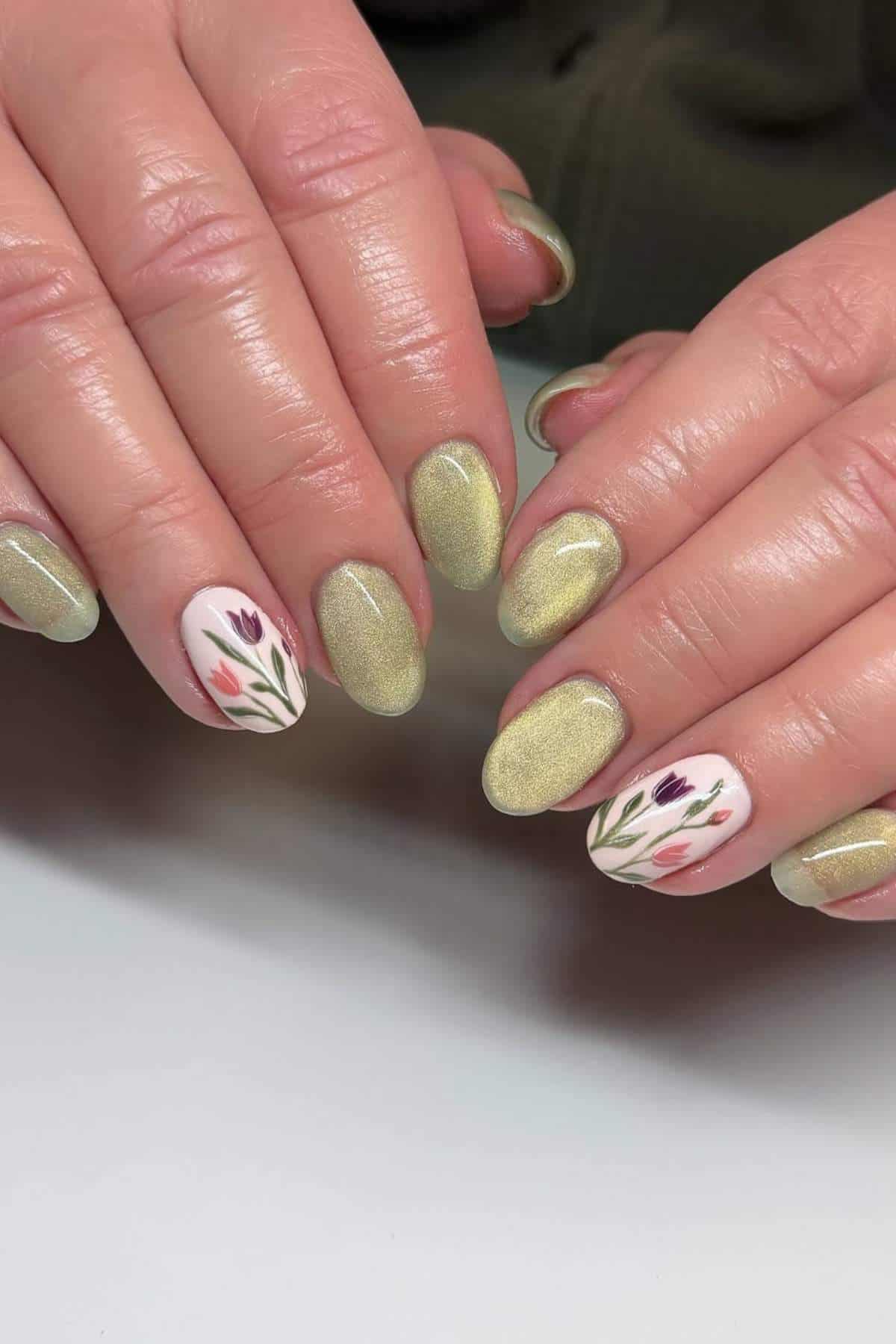 Hands with gold metallic nail polish; two accent nails feature a white base with painted pink and purple floral designs.