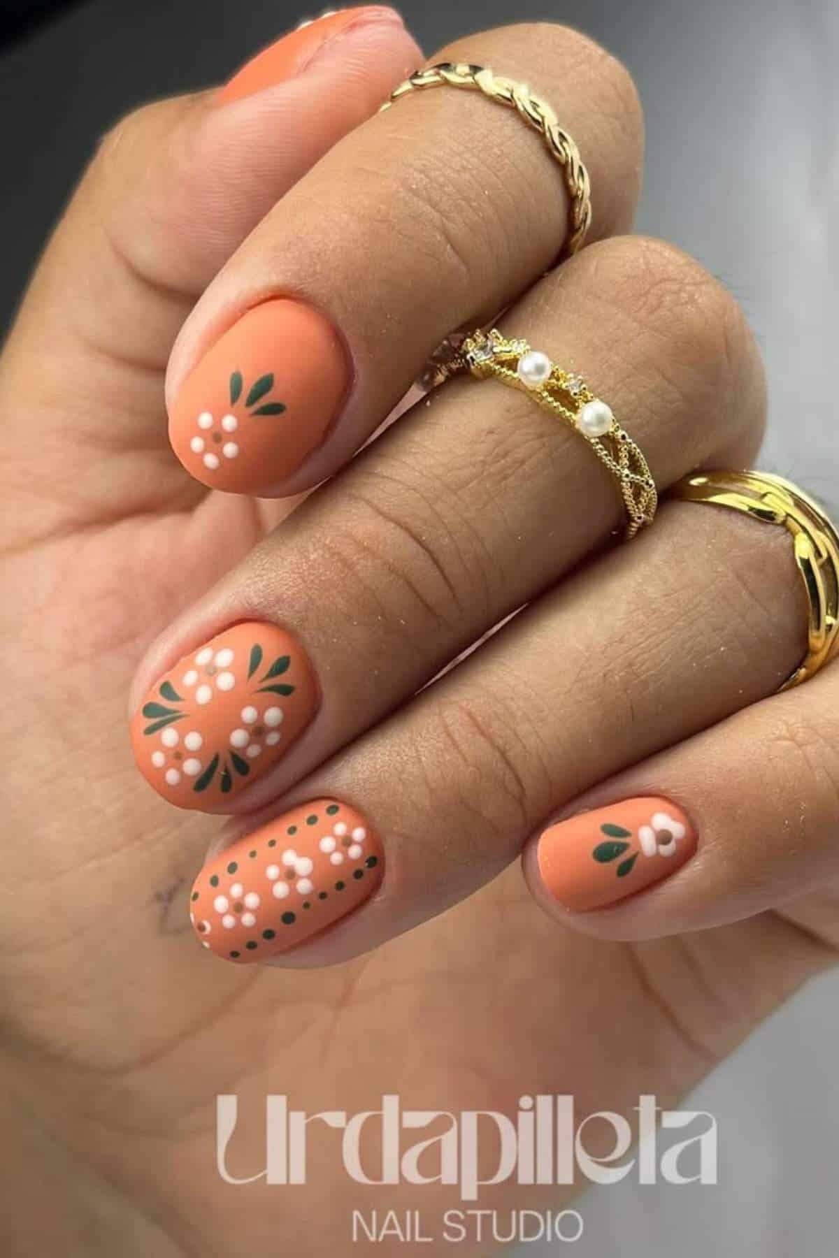 A hand with orange nail polish features white floral designs with green leaves on each fingernail. The person wears two gold rings and a gold ring with a pearl. Text reads "Urdapilleta Nail Studio.