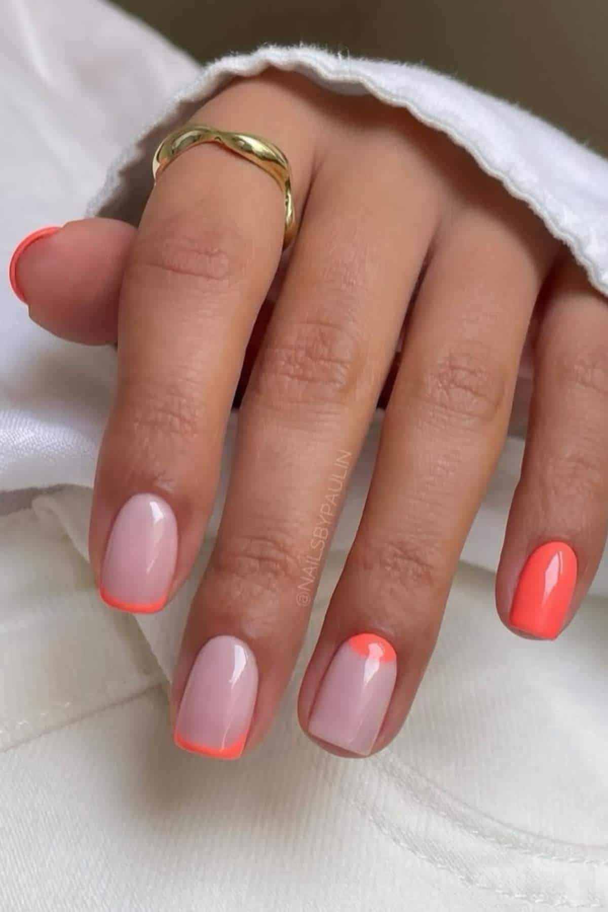 A hand with short, neatly manicured nails featuring a light pink base and bright coral French tips, wearing a gold twisted ring.