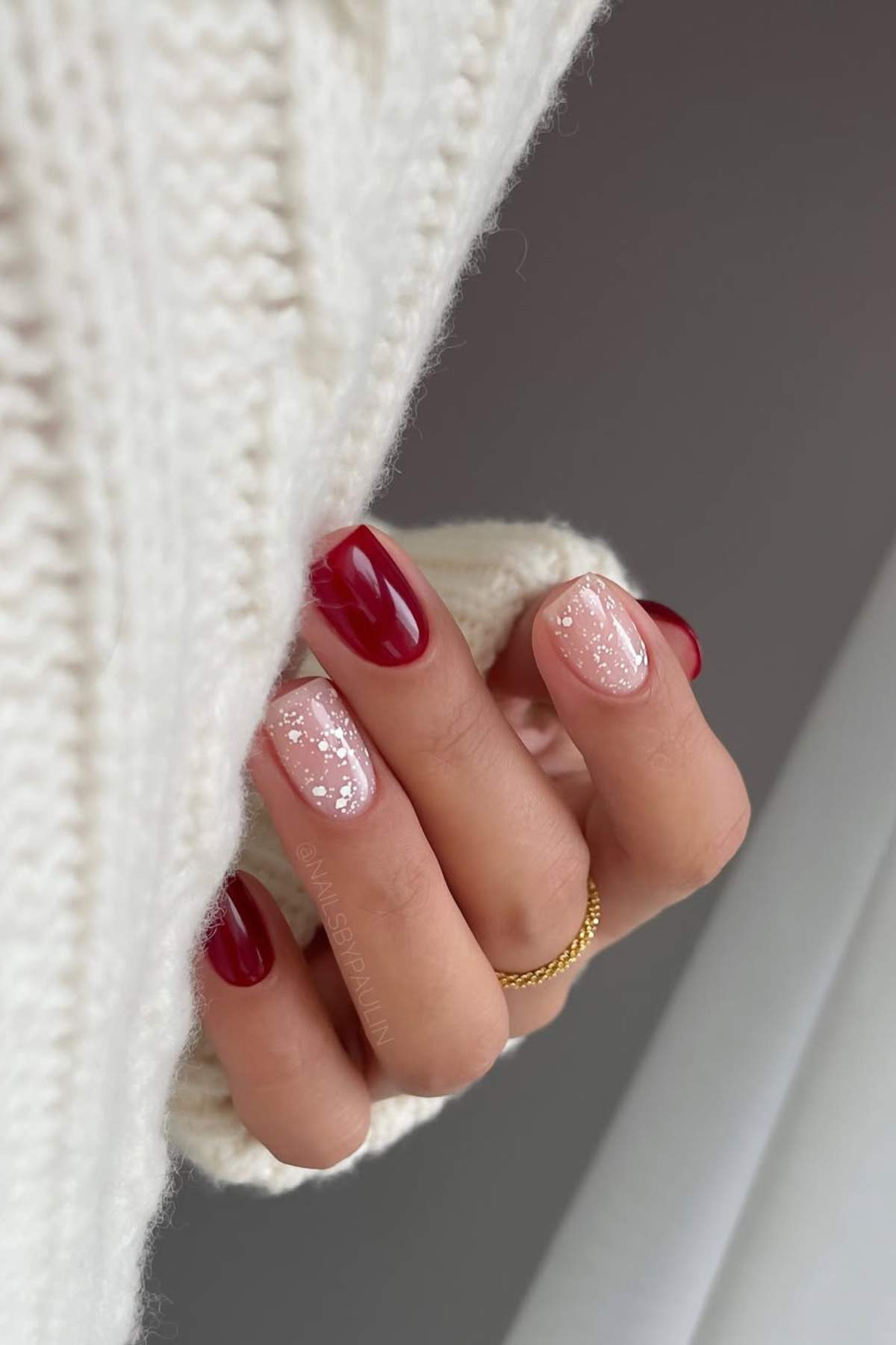 A hand with a gold ring shows nails painted red and pink with white speckles, partially covered by a white knitted sweater.