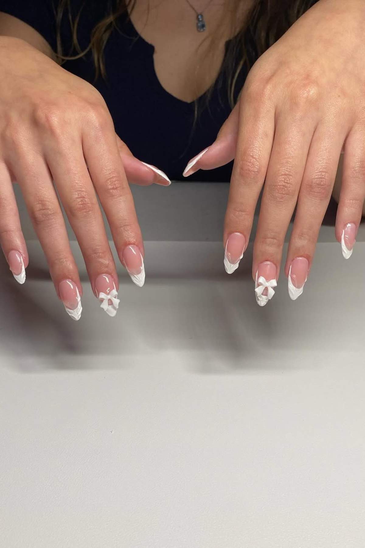 A person with long, almond-shaped nails featuring white French tips and 3D white floral designs on a few nails rests their hands on a gray surface.