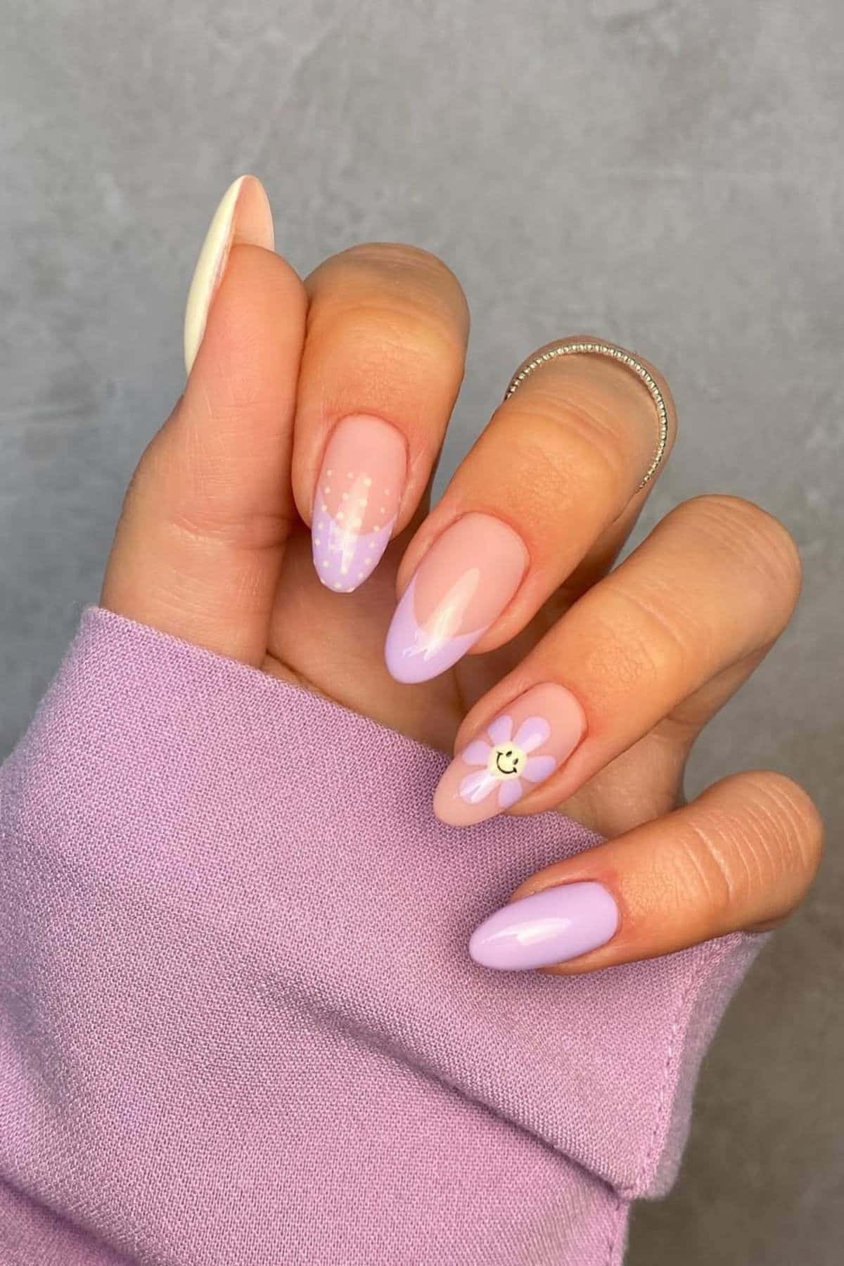 A hand with almond-shaped nails painted in pastel purple and nude shades, featuring a daisy flower design and polka dots on a few nails.