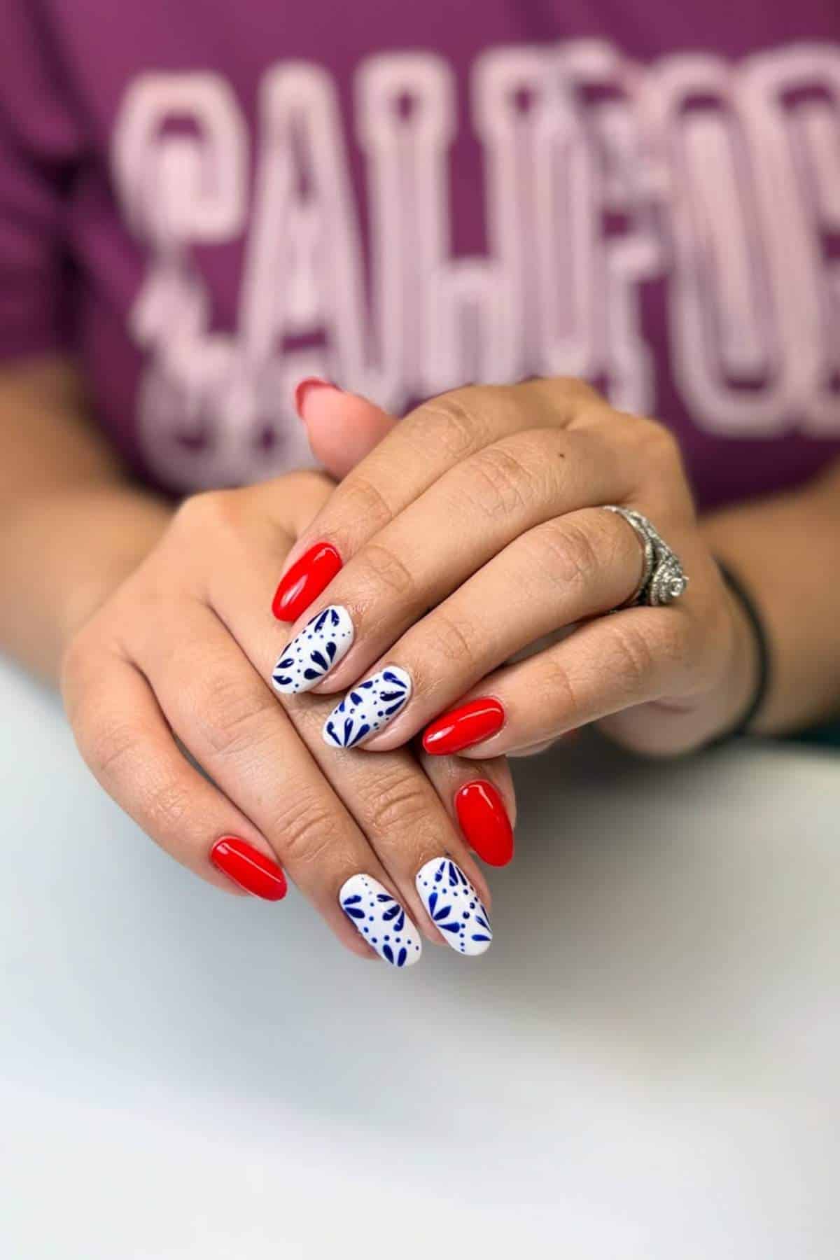 Hands with manicured nails; some nails are painted solid red, while others are white with blue leaf patterns. Person is wearing a ring and a purple "California" shirt.