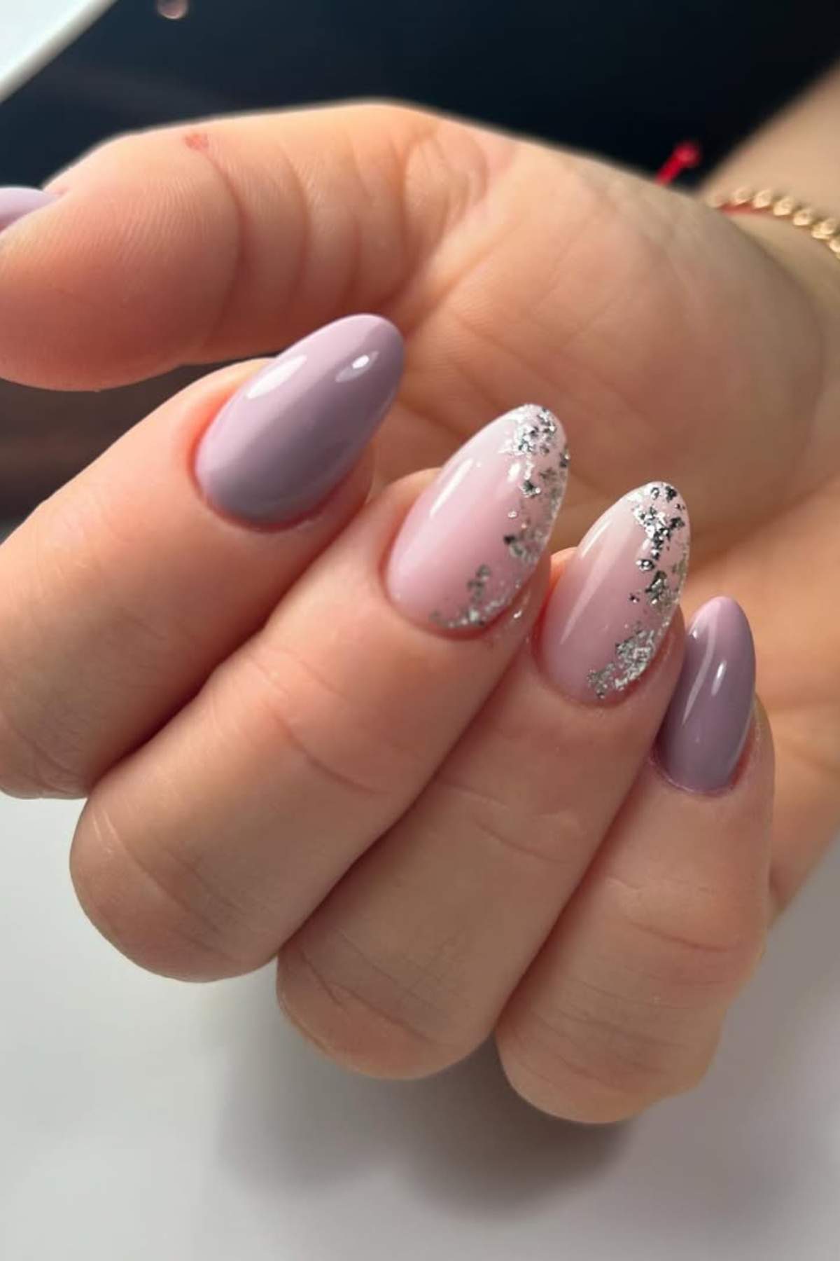 A hand with almond-shaped nails painted in light purple and pale pink, with two nails featuring silver glitter accents.