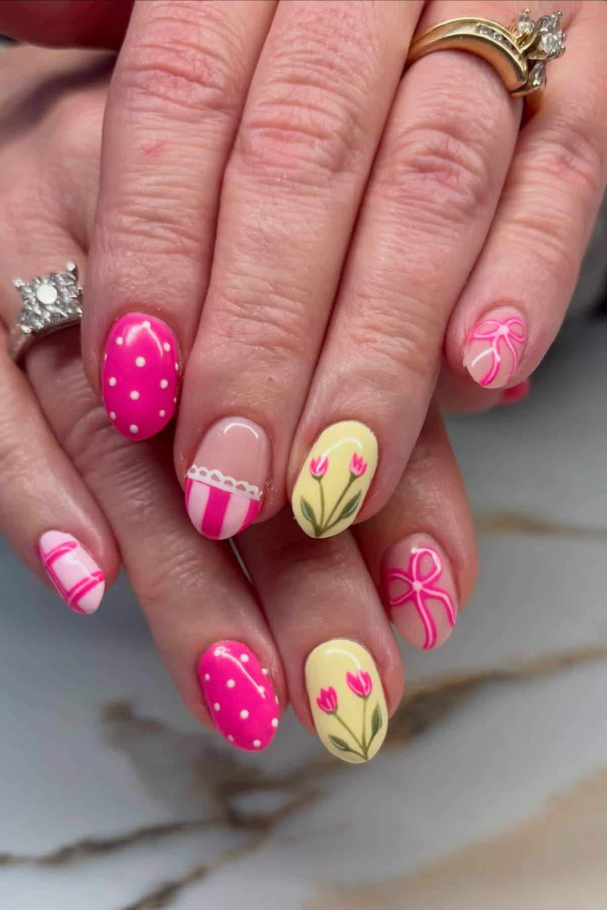Hands with manicured nails featuring pink and yellow designs, including polka dots, stripes, tulips, and ribbon patterns. The person is wearing two rings.