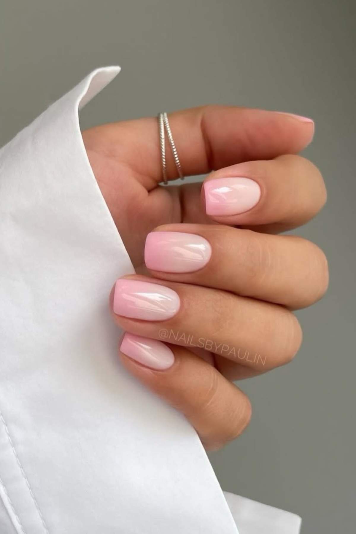 A hand with neatly manicured nails painted in a glossy light pink shade, wearing a silver ring and holding the cuff of a white shirt.