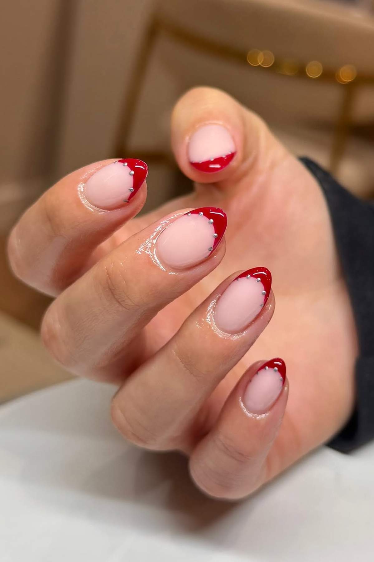 A hand with almond-shaped nails featuring a nude base and glossy red, irregular French tips.