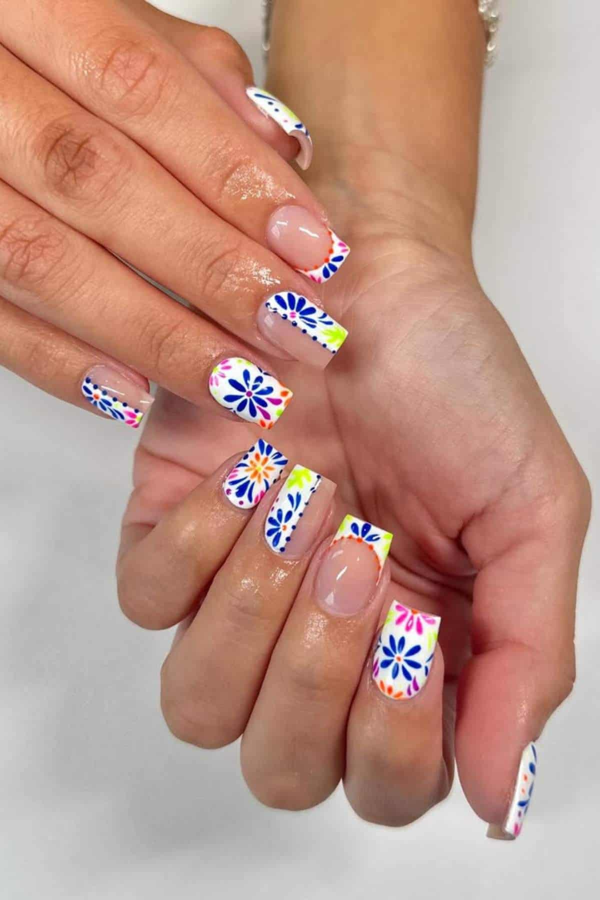 A person’s hands with square-shaped nails featuring white tips and colorful floral designs on each nail.