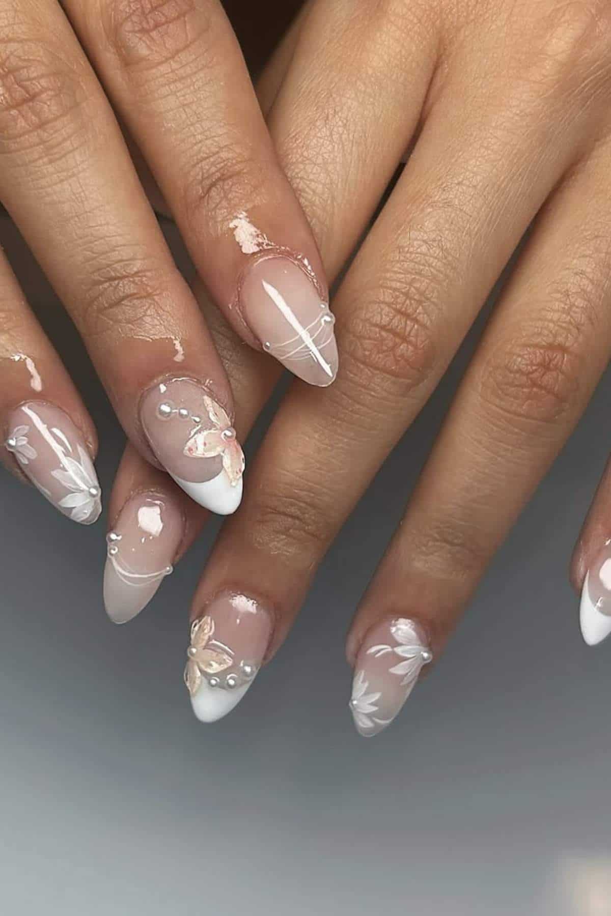 Hands with almond-shaped nails featuring French tips, white floral designs, and small pearl embellishments on a neutral background.