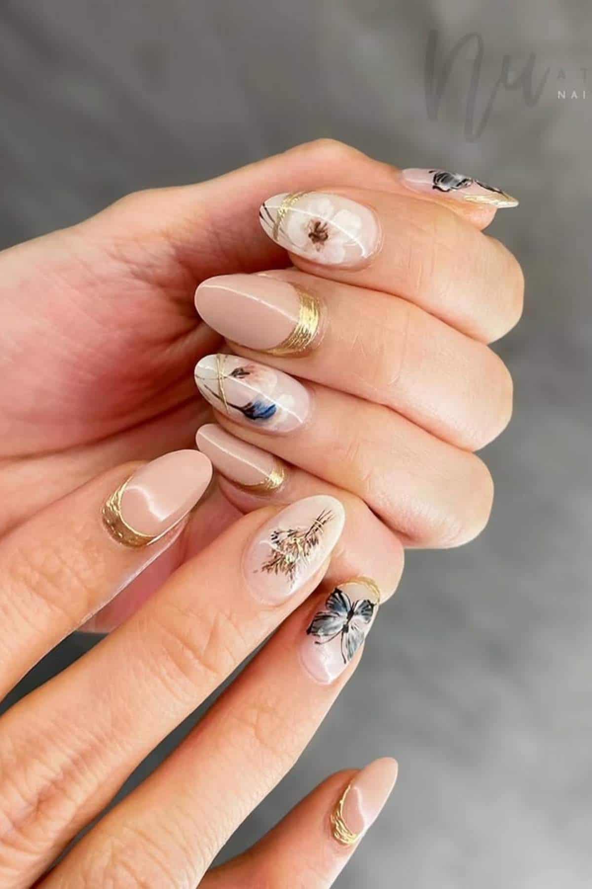 A hand with almond-shaped nails featuring nude polish, gold accents, and delicate floral designs on some nails.