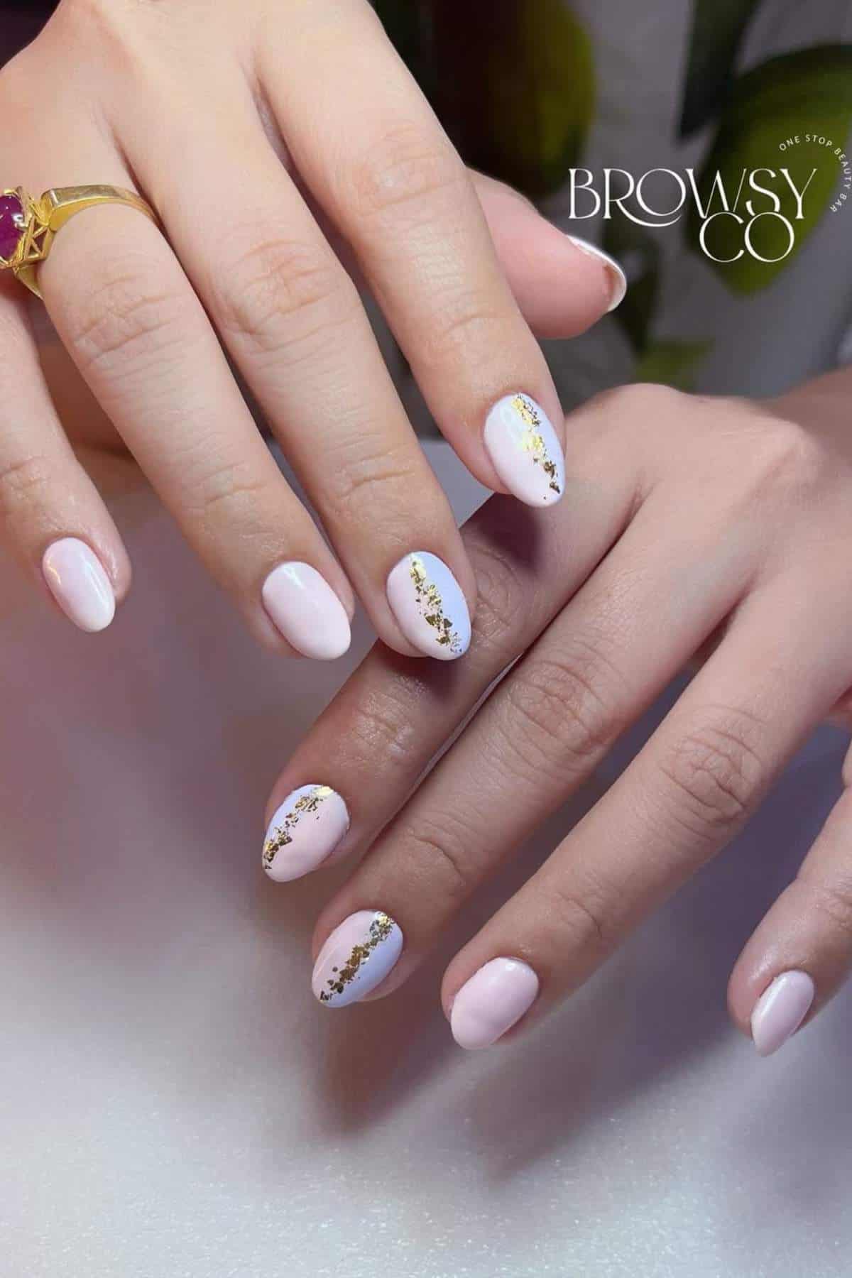 Close-up of hands with light pink manicured nails, featuring gold foil accents on some nails. A gold ring with a purple stone is worn on one finger.