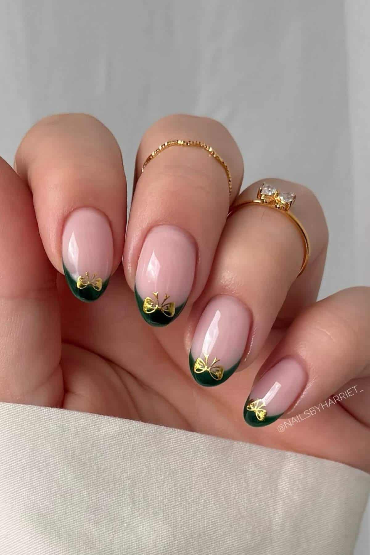 A hand with almond-shaped nails featuring nude polish, dark green French tips, and gold bow nail art; two gold rings are worn on the fingers.