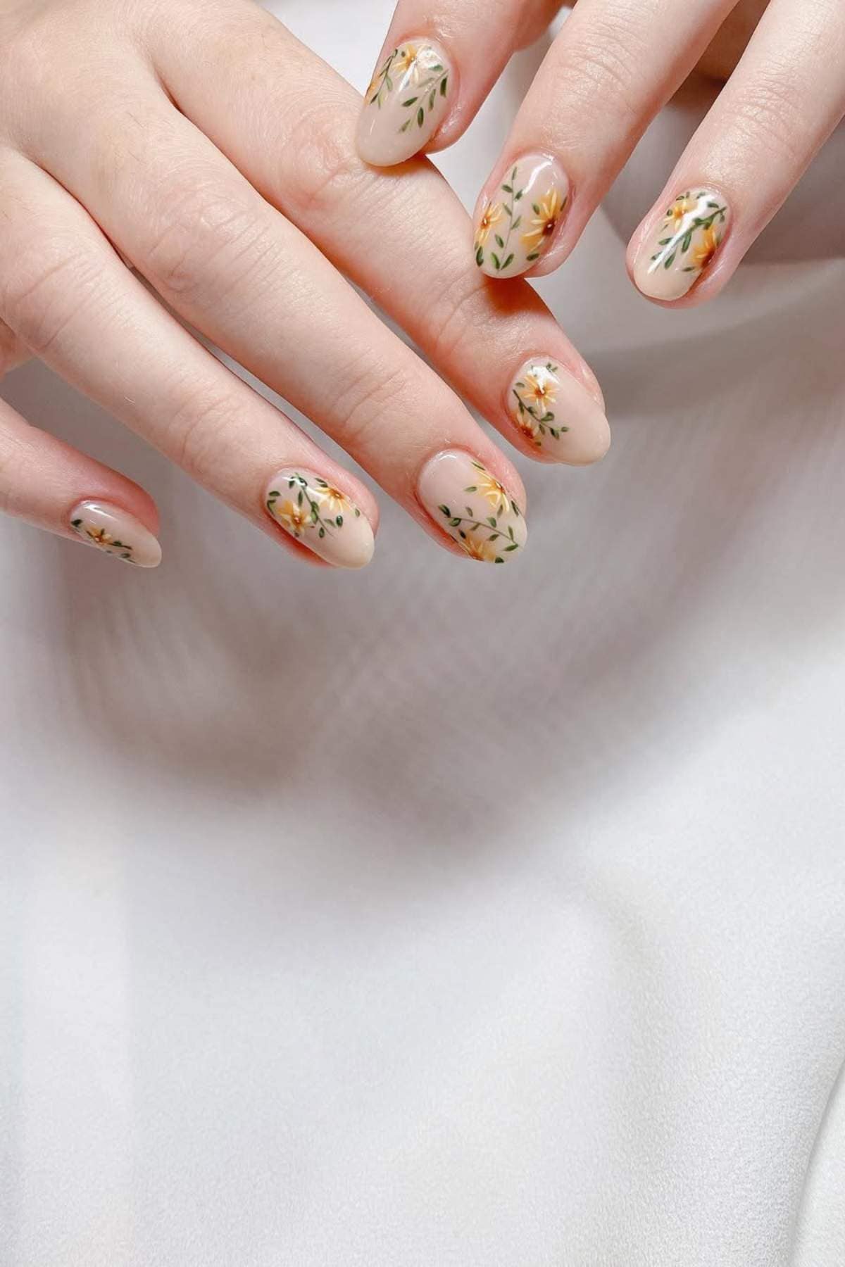 Hands with almond-shaped nails featuring a light nude base and delicate yellow and orange floral designs on each nail.