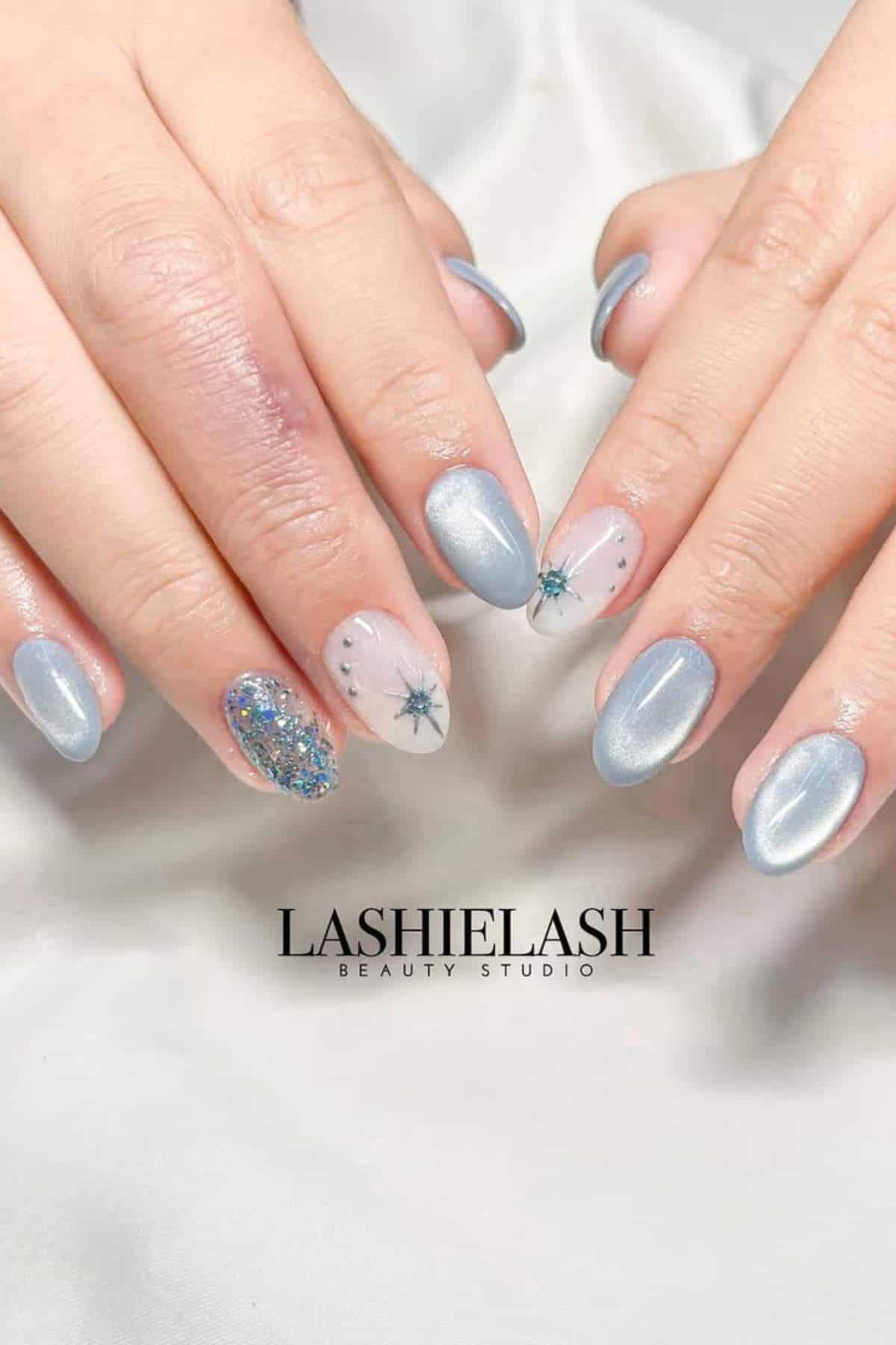 Hands with oval-shaped, silver metallic nails featuring accent designs: glitter on one nail and starburst patterns on two nails, displayed on a white surface with "LASHIELASH" text.