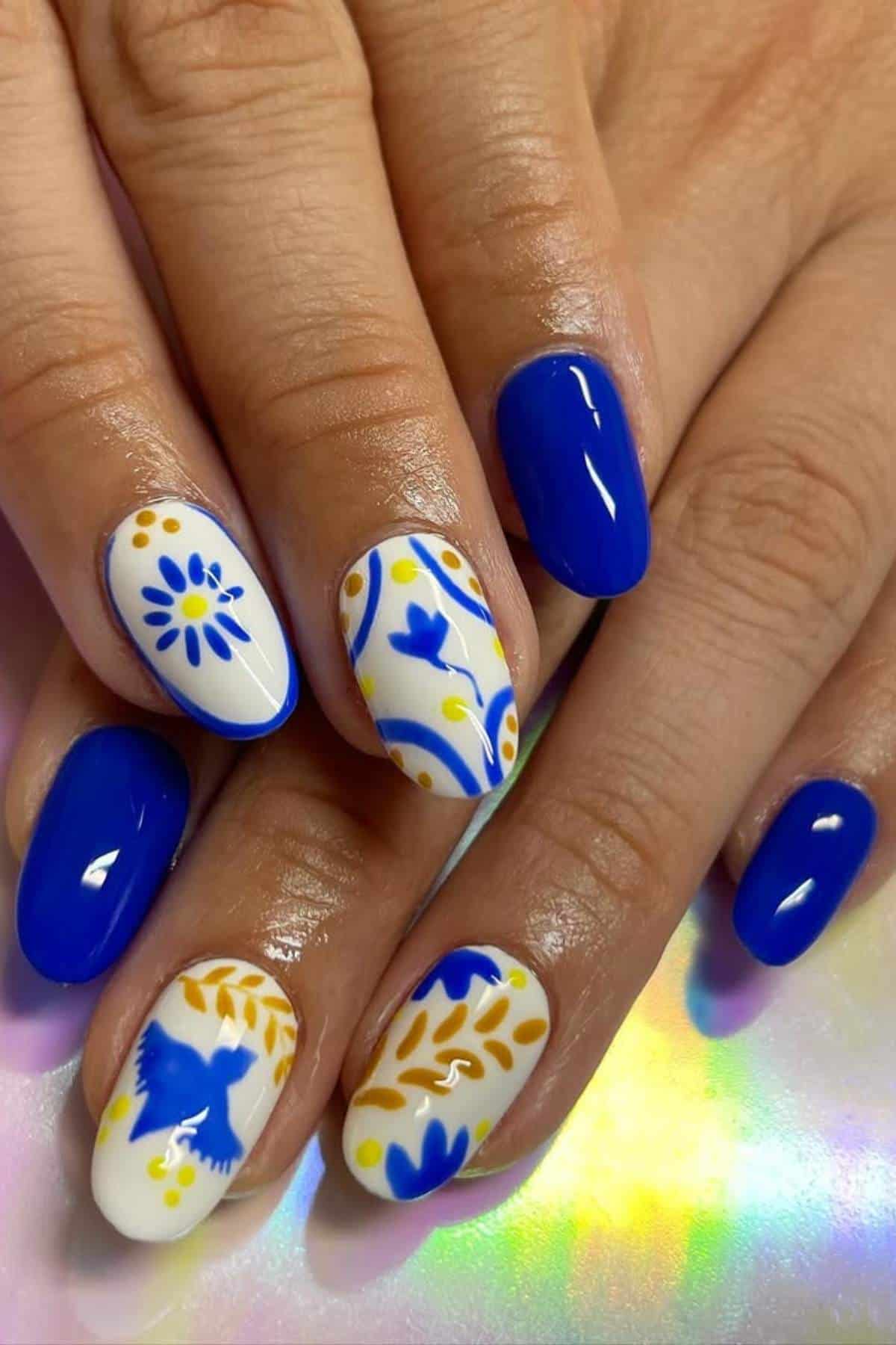 Hands with blue, white, and yellow nail art; some nails are solid blue while others feature floral and bird designs on a white background.