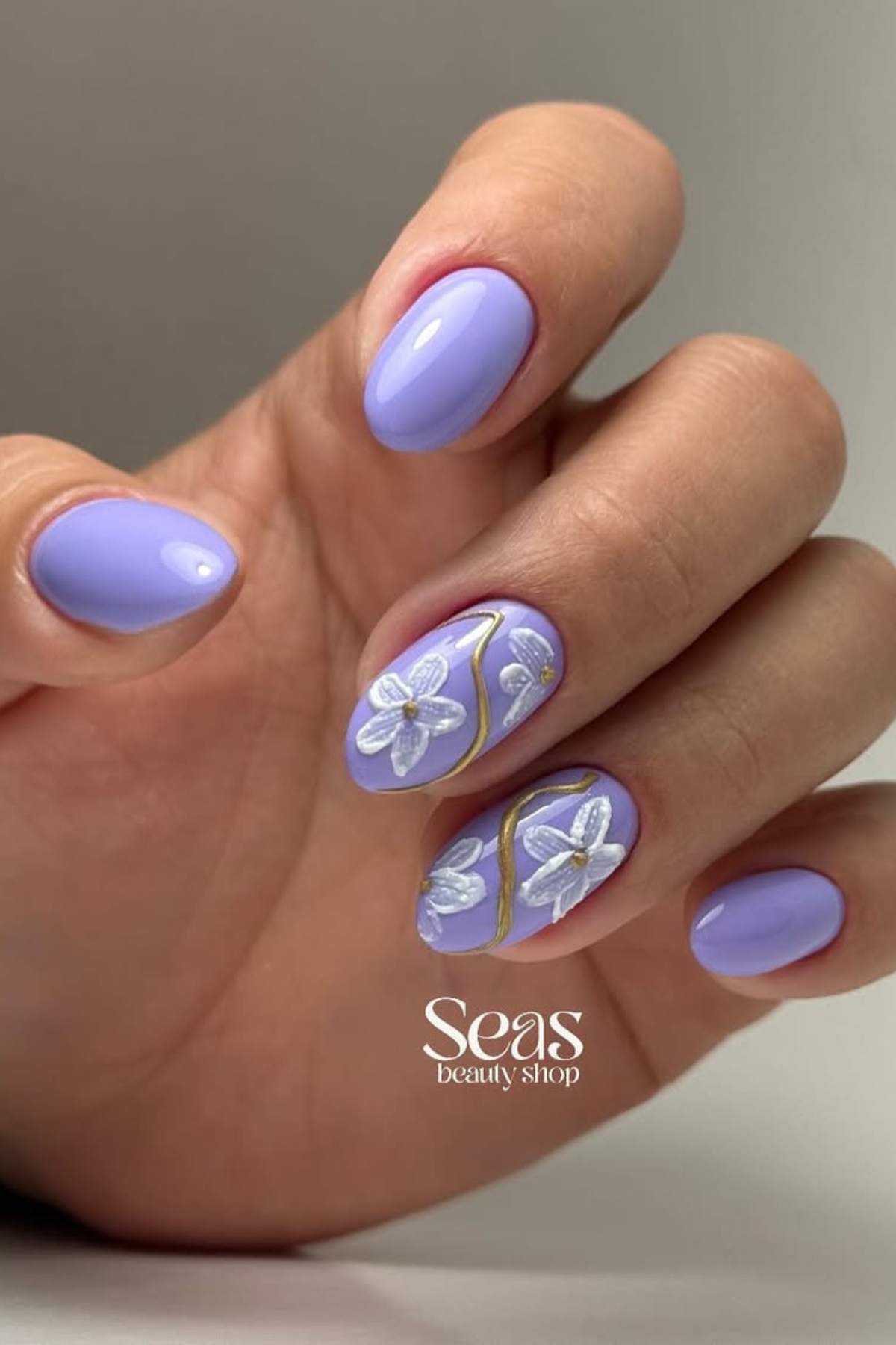 A hand with lavender-colored nails, two accent nails featuring white 3D floral designs with gold lines, and the words "Seas beauty shop" in the corner.