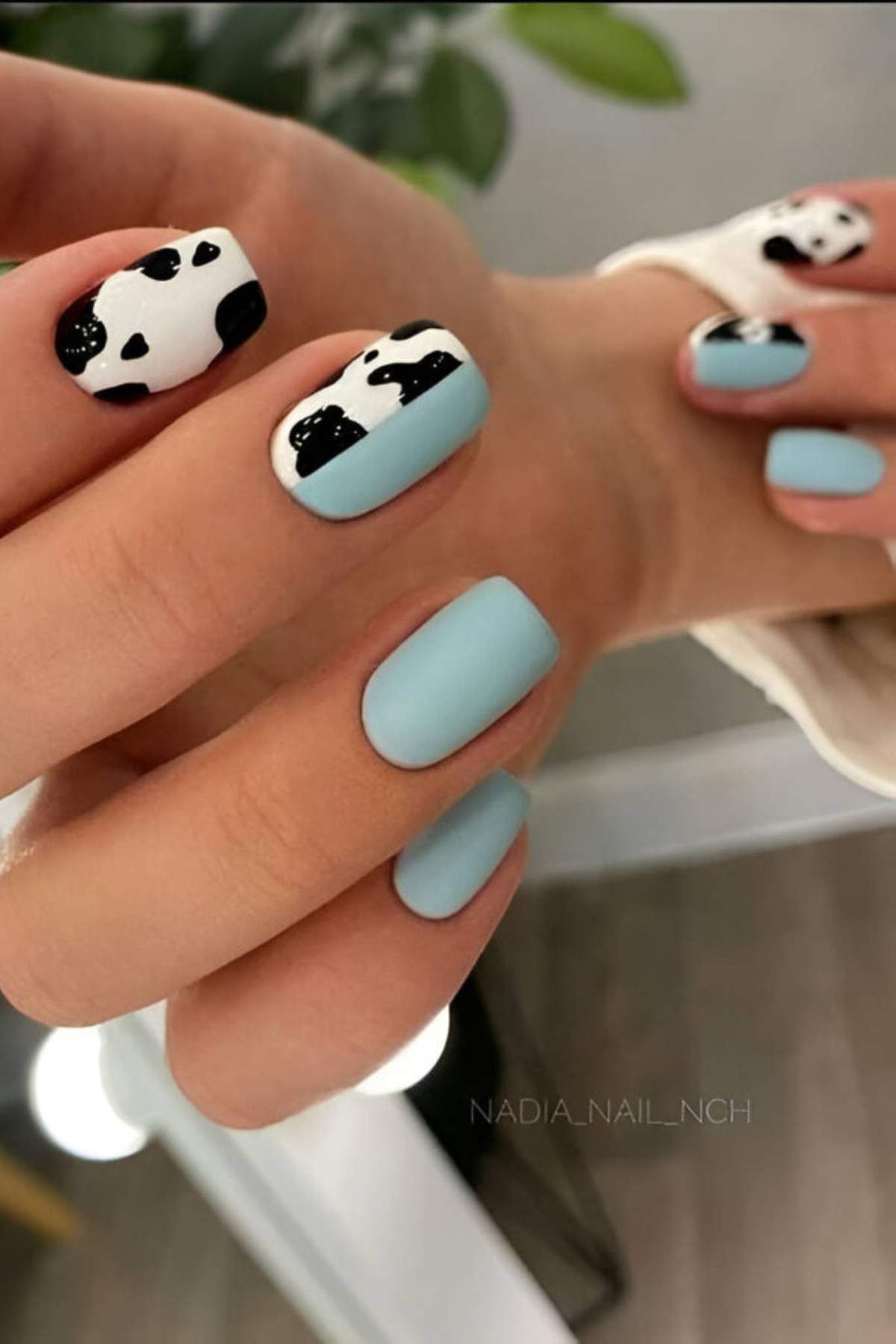 Close-up of hands with manicured nails; most nails are painted light blue, while the index and middle fingernails have a black and white cow print design.