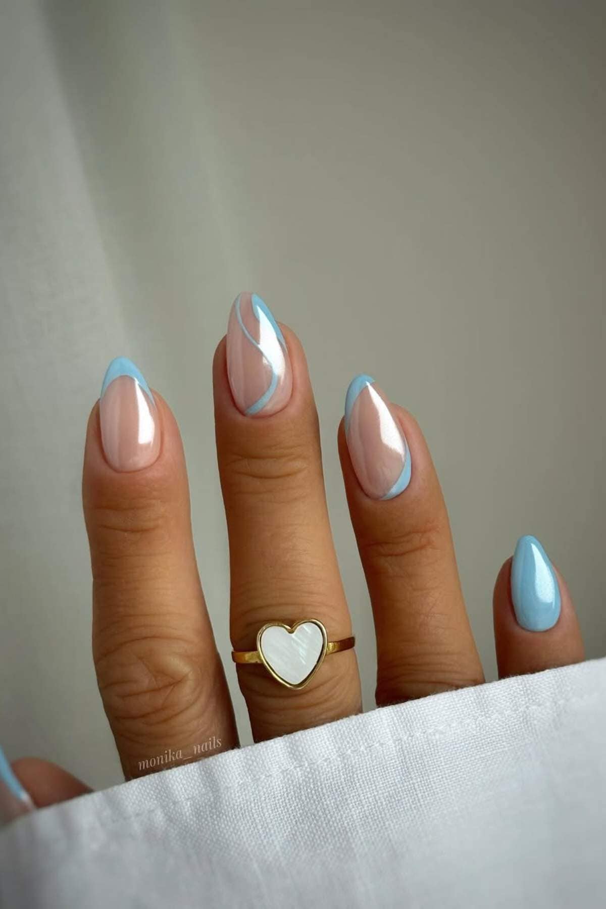 A hand with light blue and nude almond-shaped nails, featuring blue French tips and abstract designs, wears a gold ring with a white heart charm.